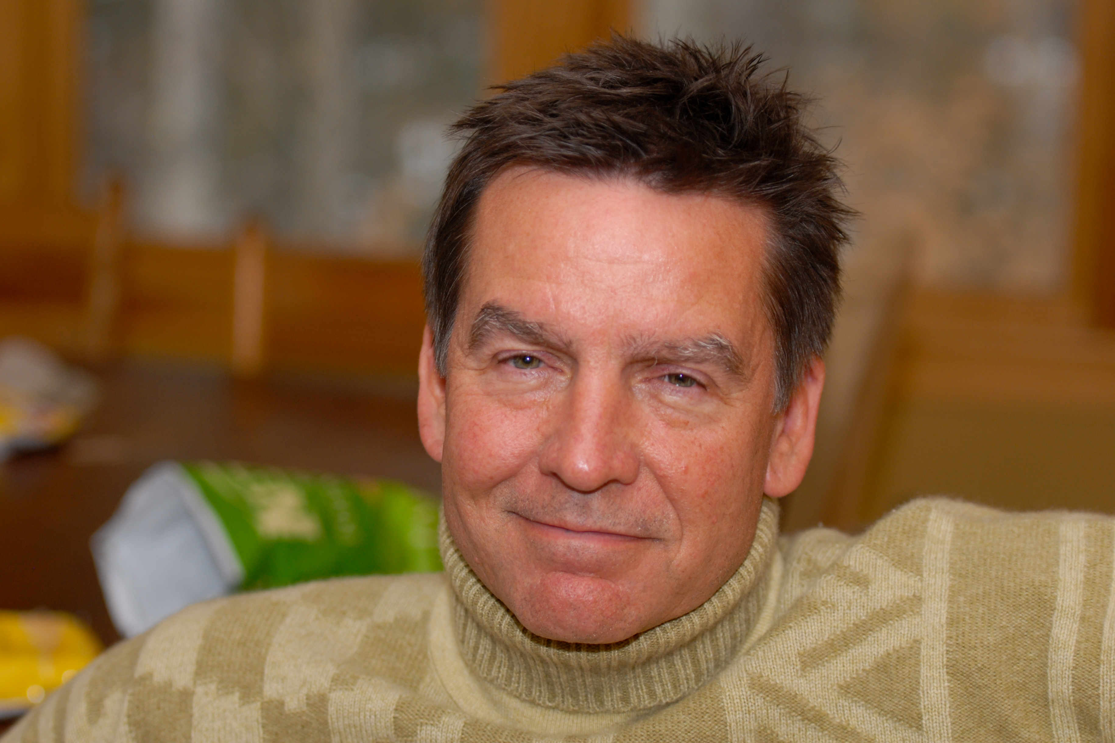 A man in a green turtleneck sweater sits indoors, looking directly at the camera with a relaxed expression.