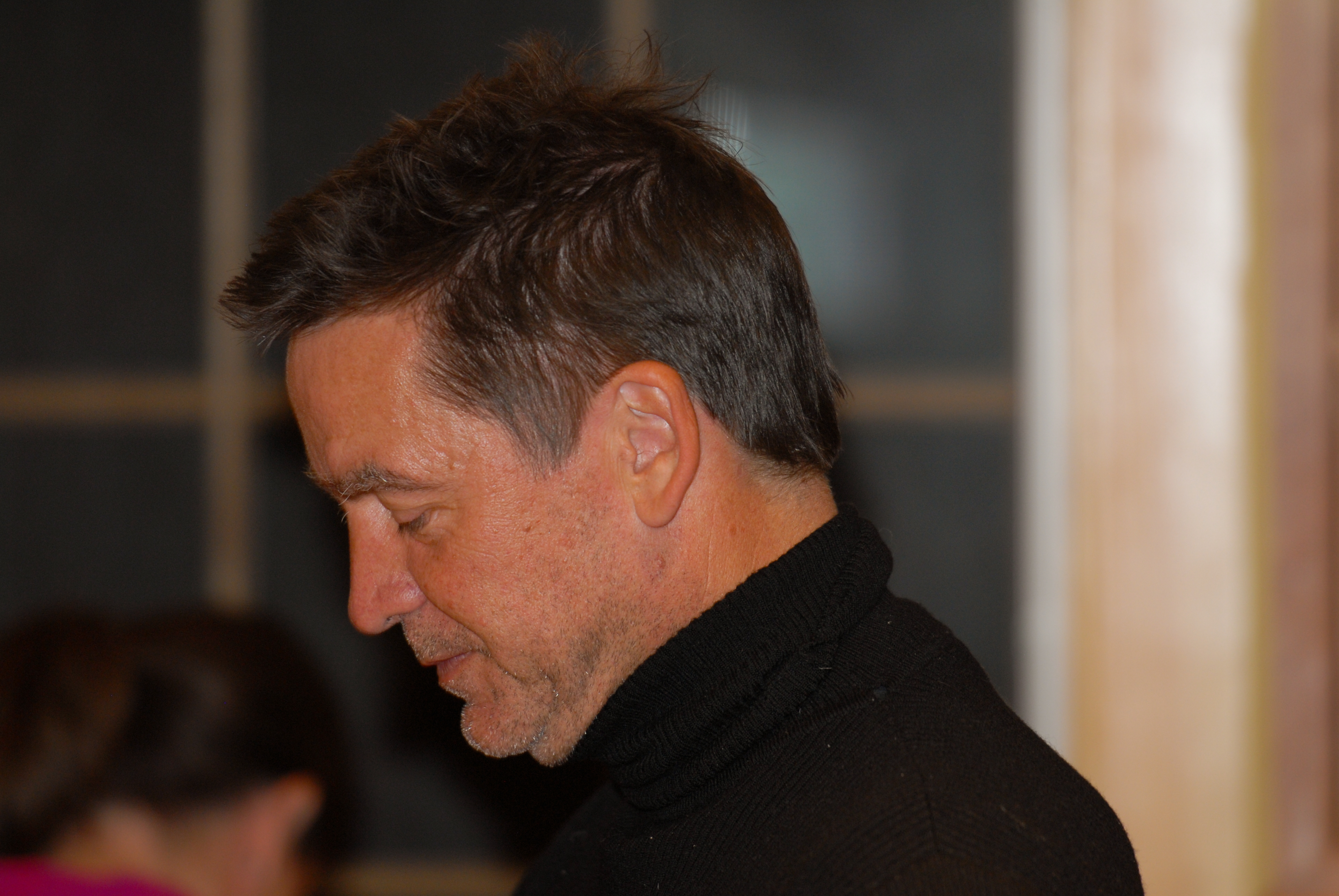 A man in a black turtleneck looks downward with a focused expression in an indoor setting.