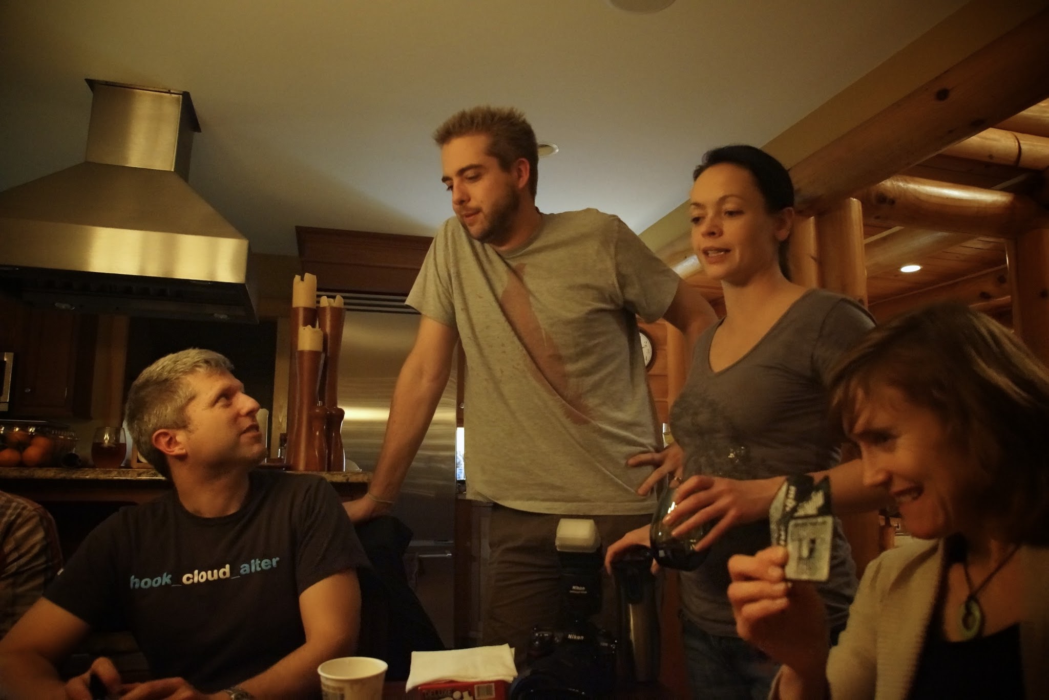 A group of people in a cabin-style kitchen, engaged in conversation and holding drinks and a card.