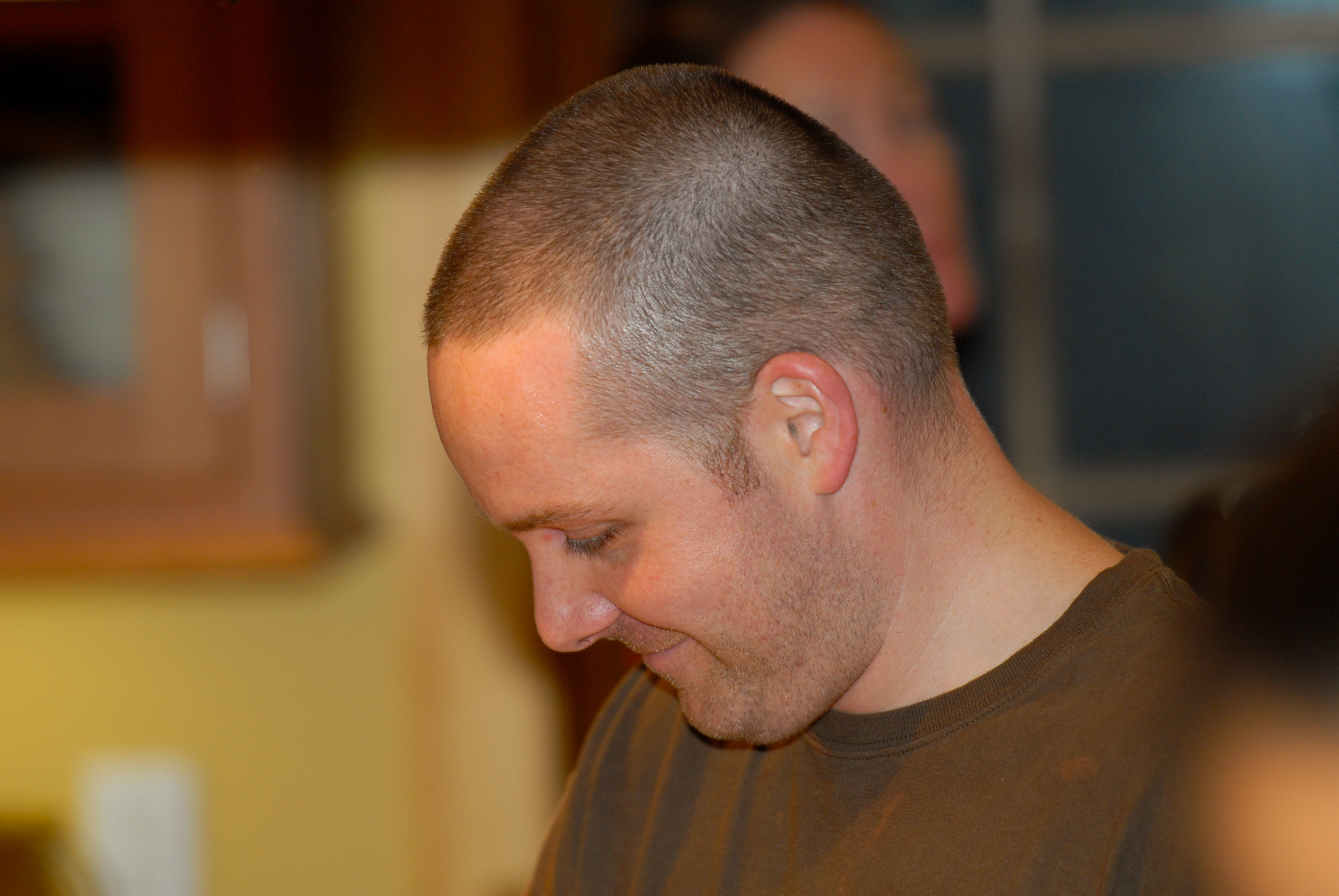 A man with a shaved head and brown shirt looks down and smiles in a warmly lit indoor setting.
