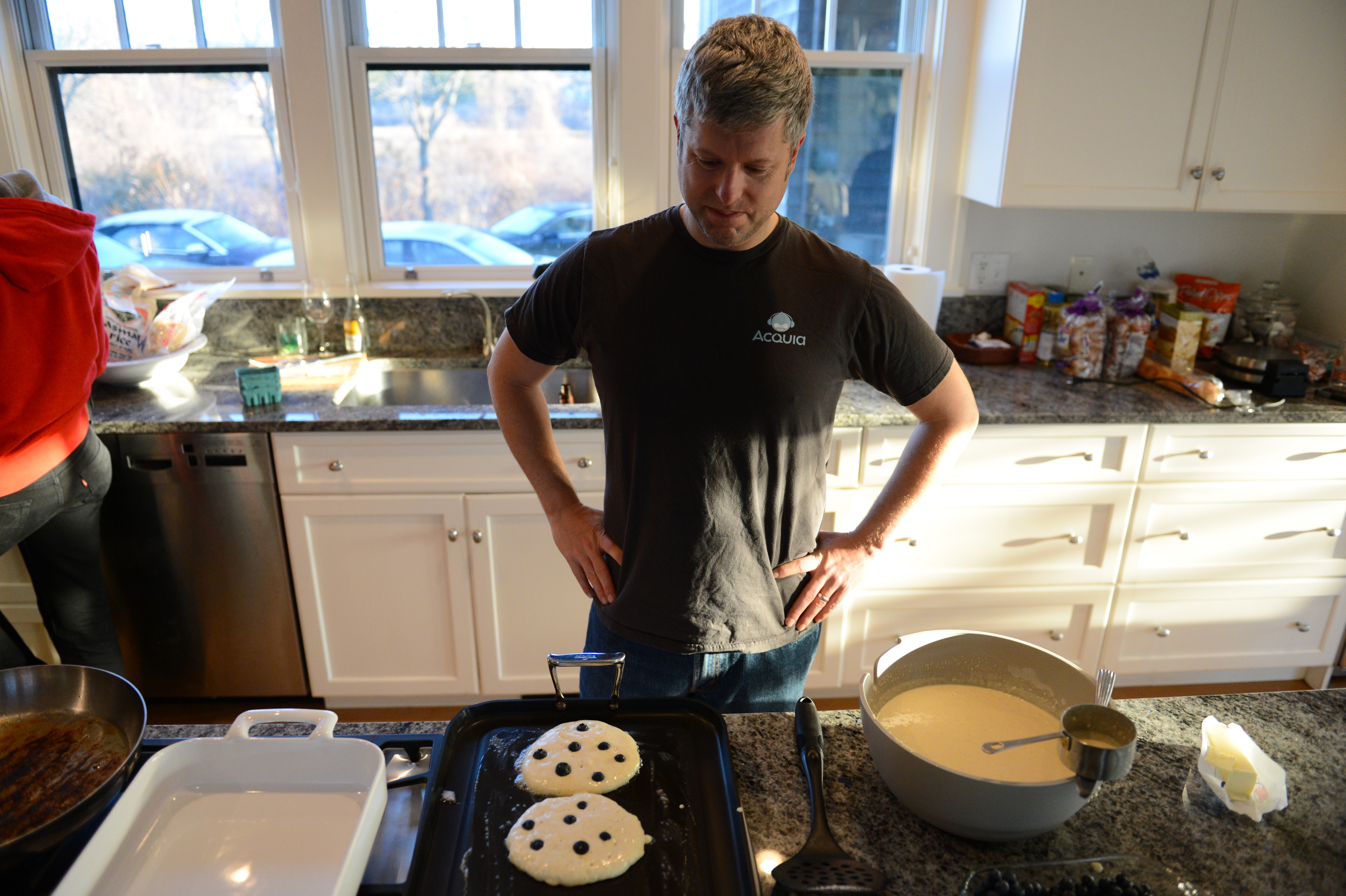 A man in an Acquia shirt looks at blueberry pancakes cooking on a griddle in a kitchen.