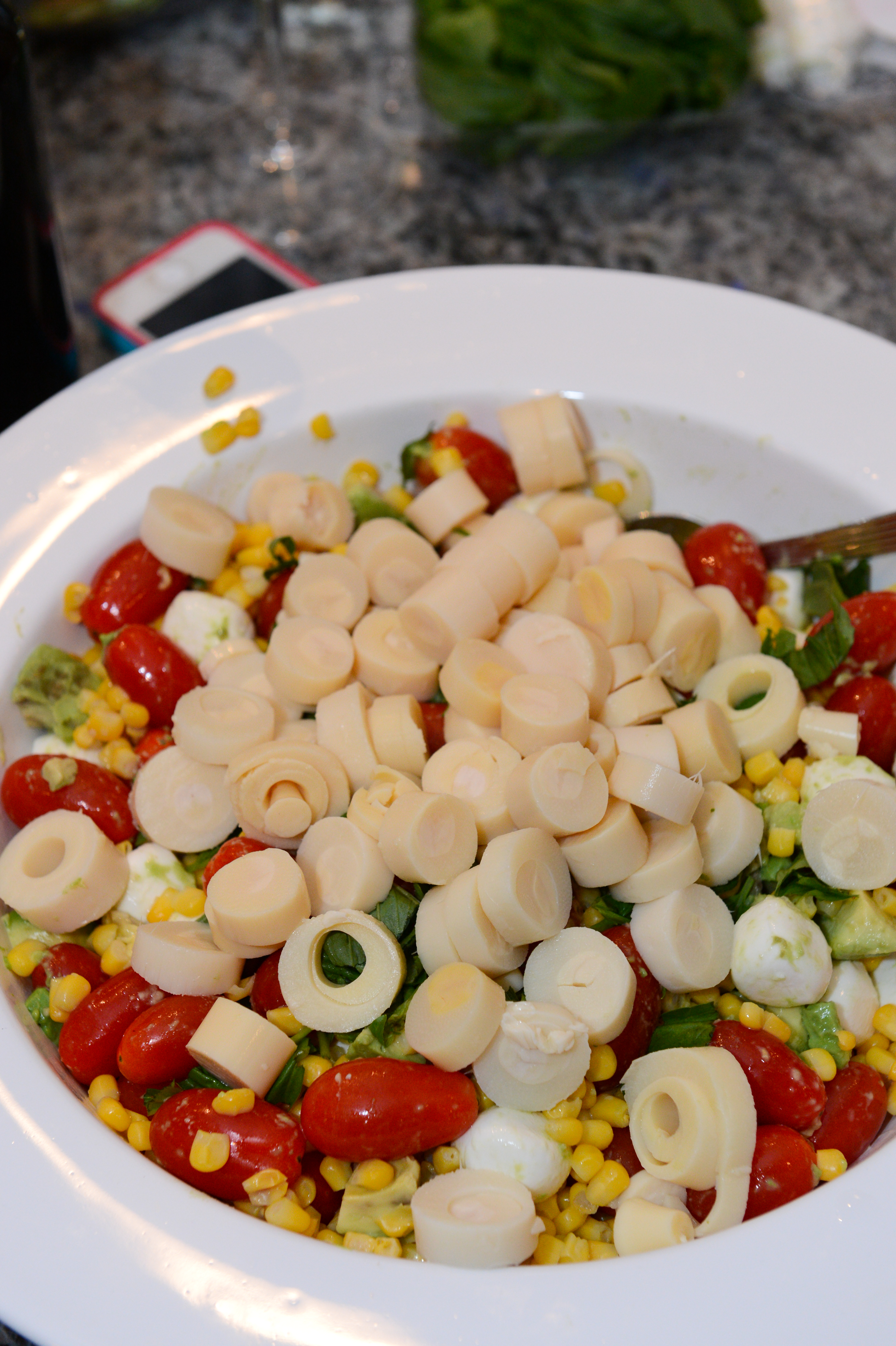A white bowl filled with salad containing cherry tomatoes, corn, mozzarella, avocado, and sliced hearts of palm.