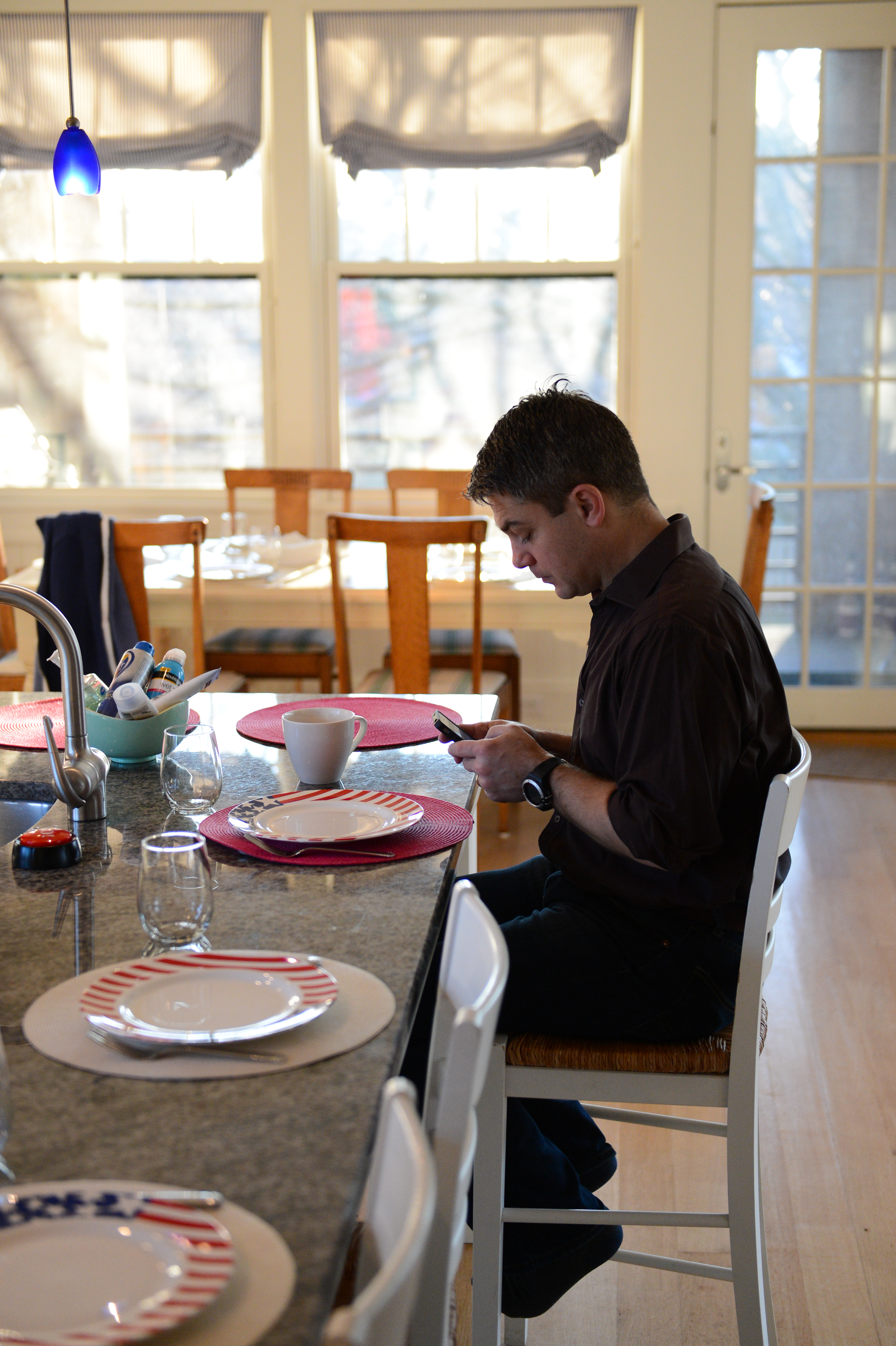A man sits at a kitchen counter, looking at his phone, with table settings and a coffee cup nearby.