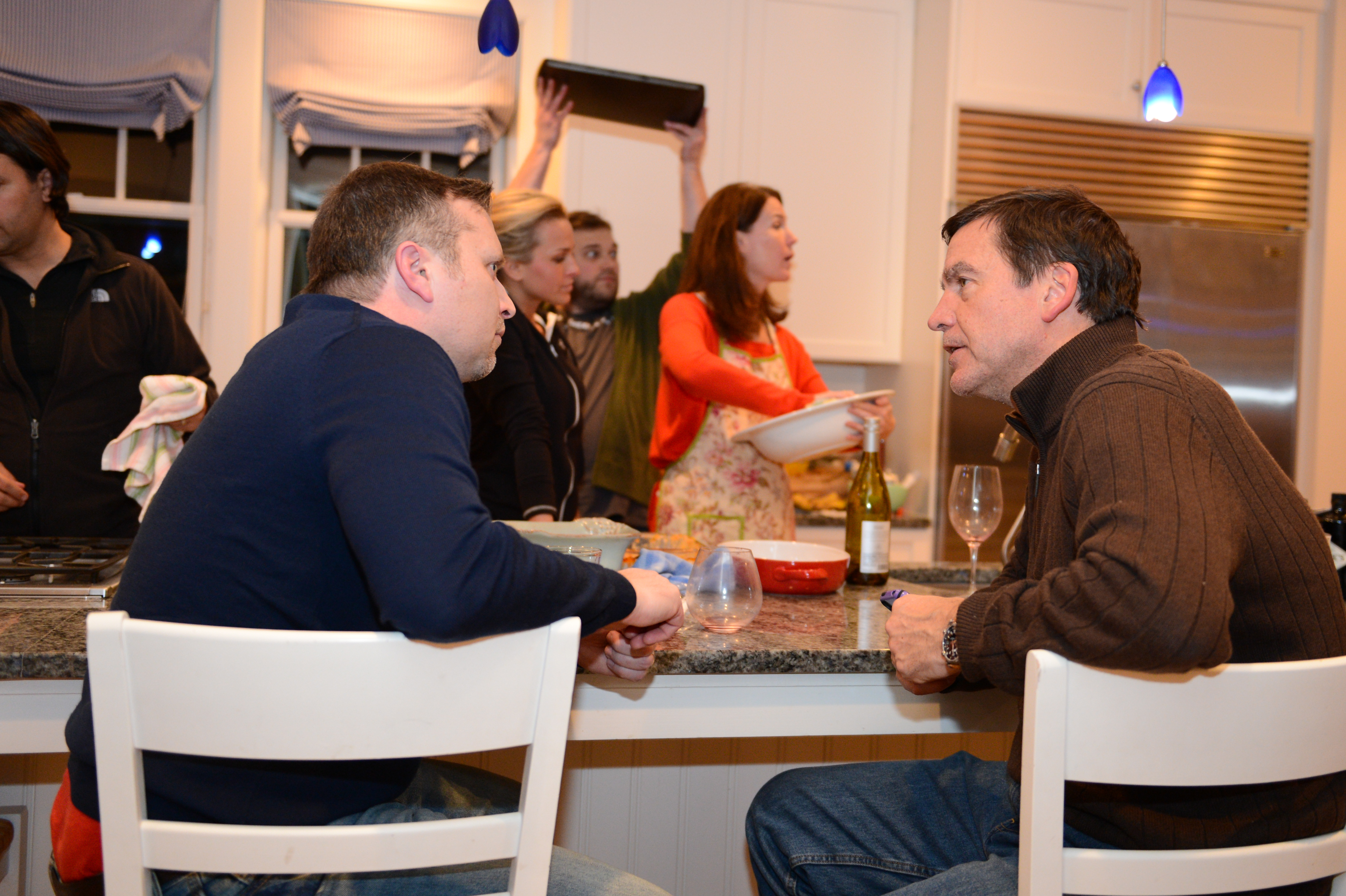 Two men sit at a kitchen counter, engaged in conversation, while others prepare food and interact in the background.