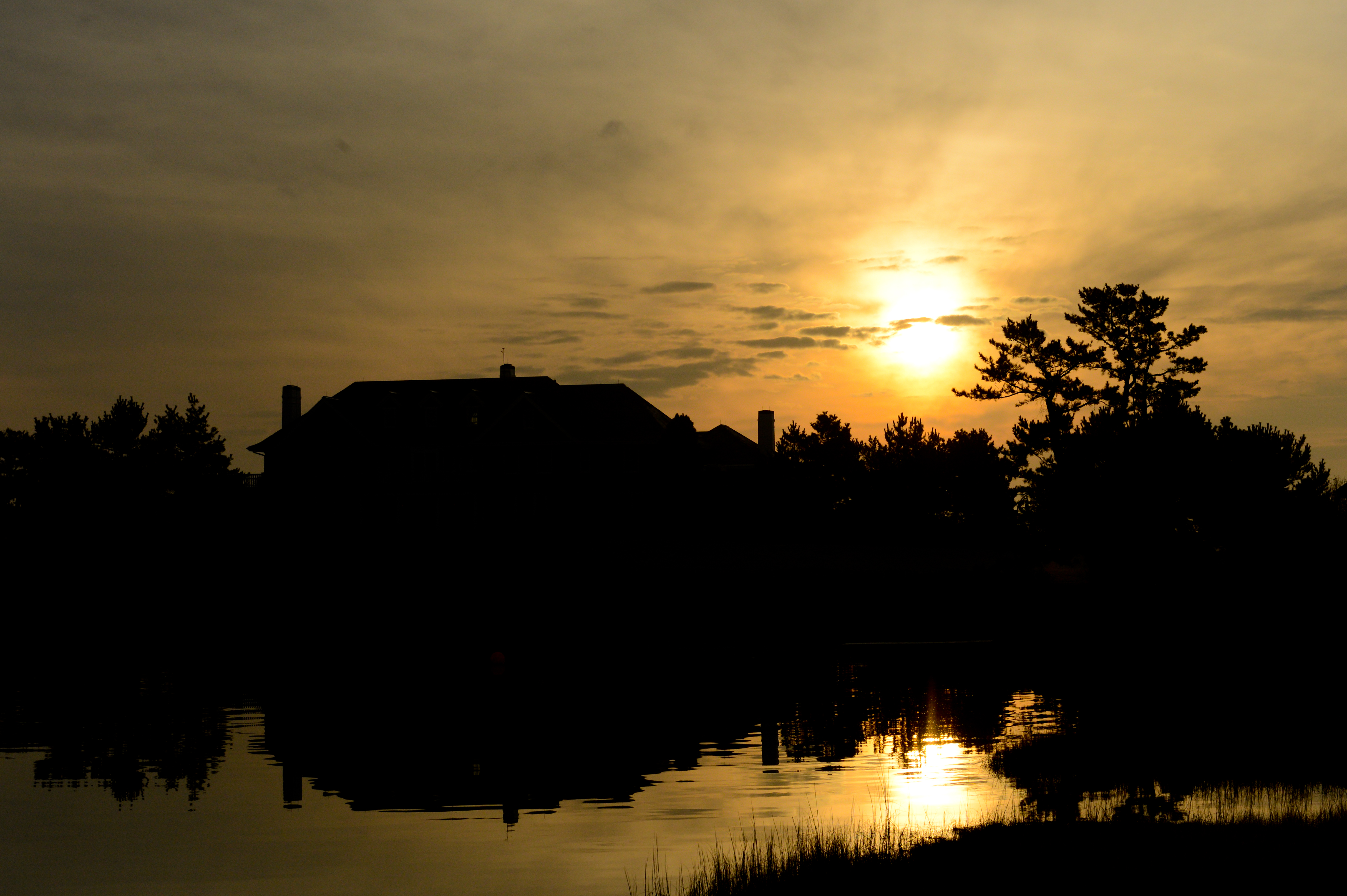 Silhouetted house and trees reflected in water at sunset, with a golden sky and scattered clouds.