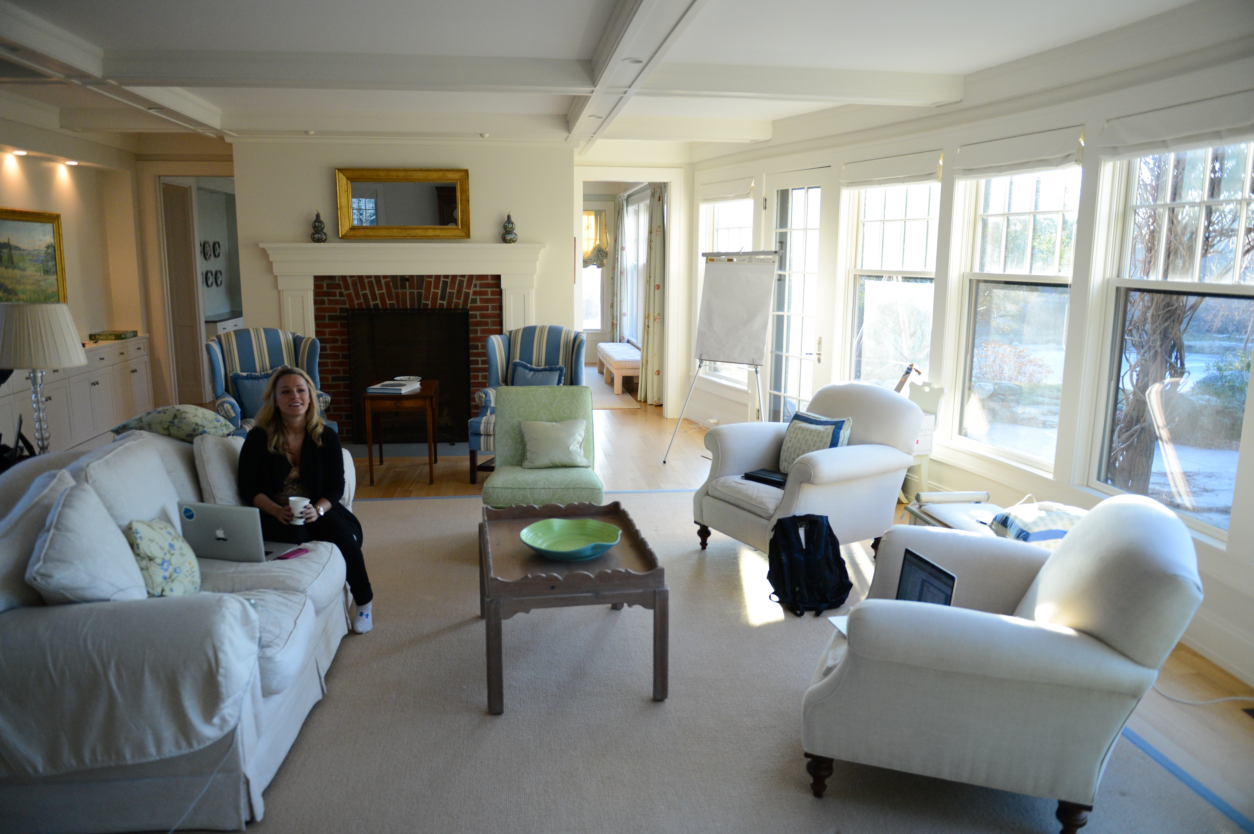 A woman sits on a couch with a laptop, holding a cup, in a bright living room.