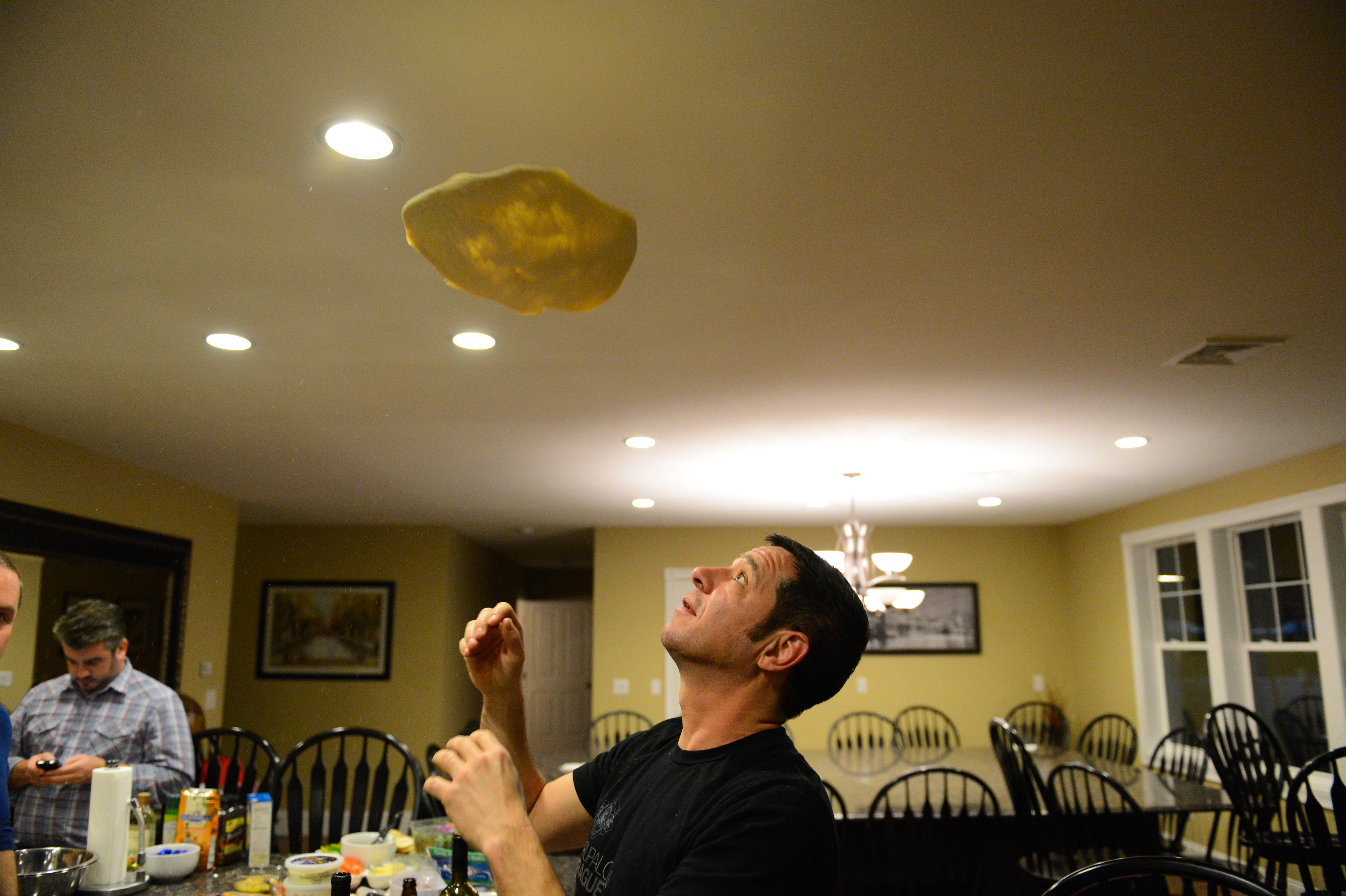 A man tosses pizza dough into the air while preparing food in a kitchen with others nearby.