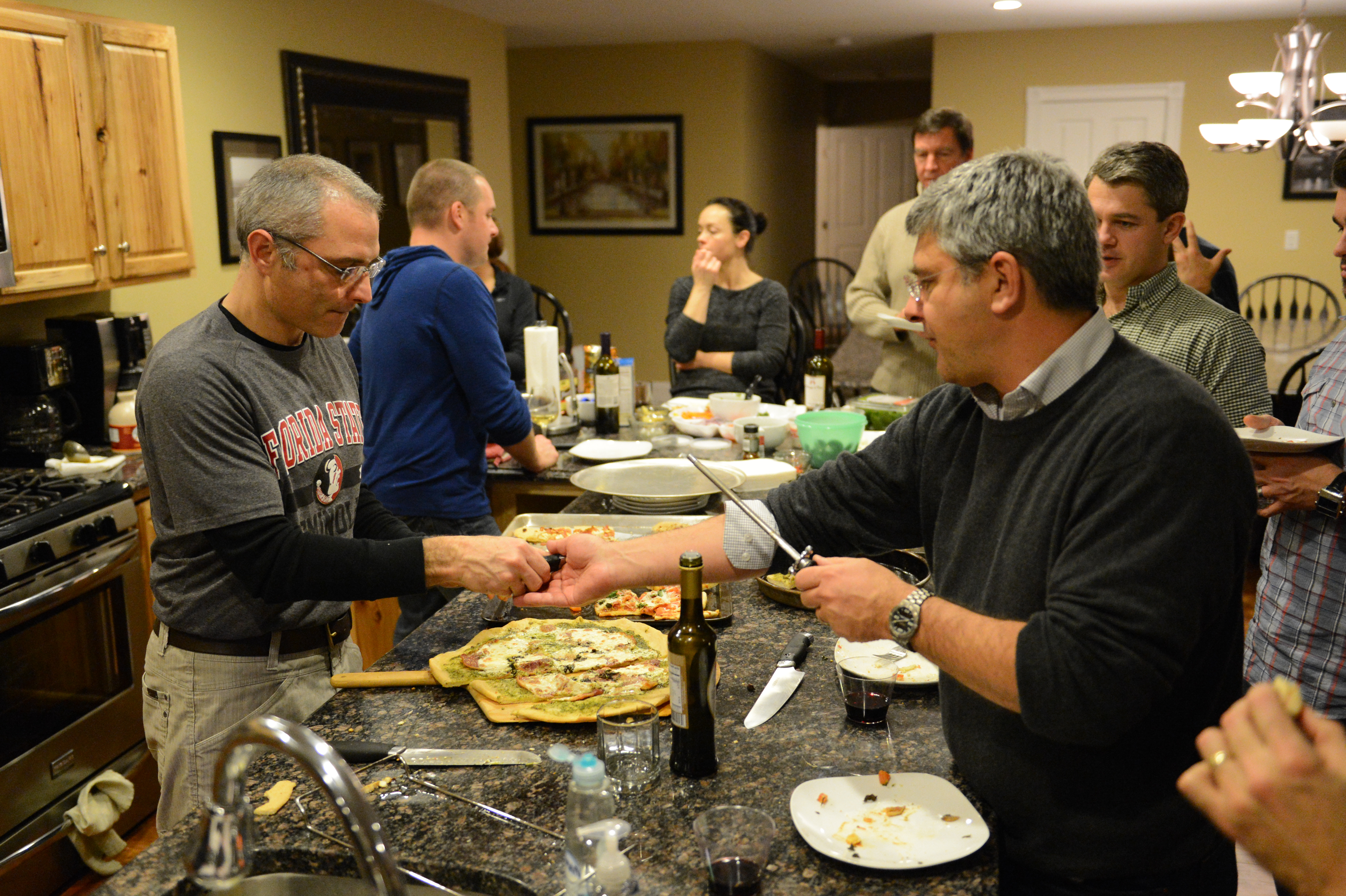 A group of people gathers in a kitchen, sharing homemade pizza and drinks during a casual social event.