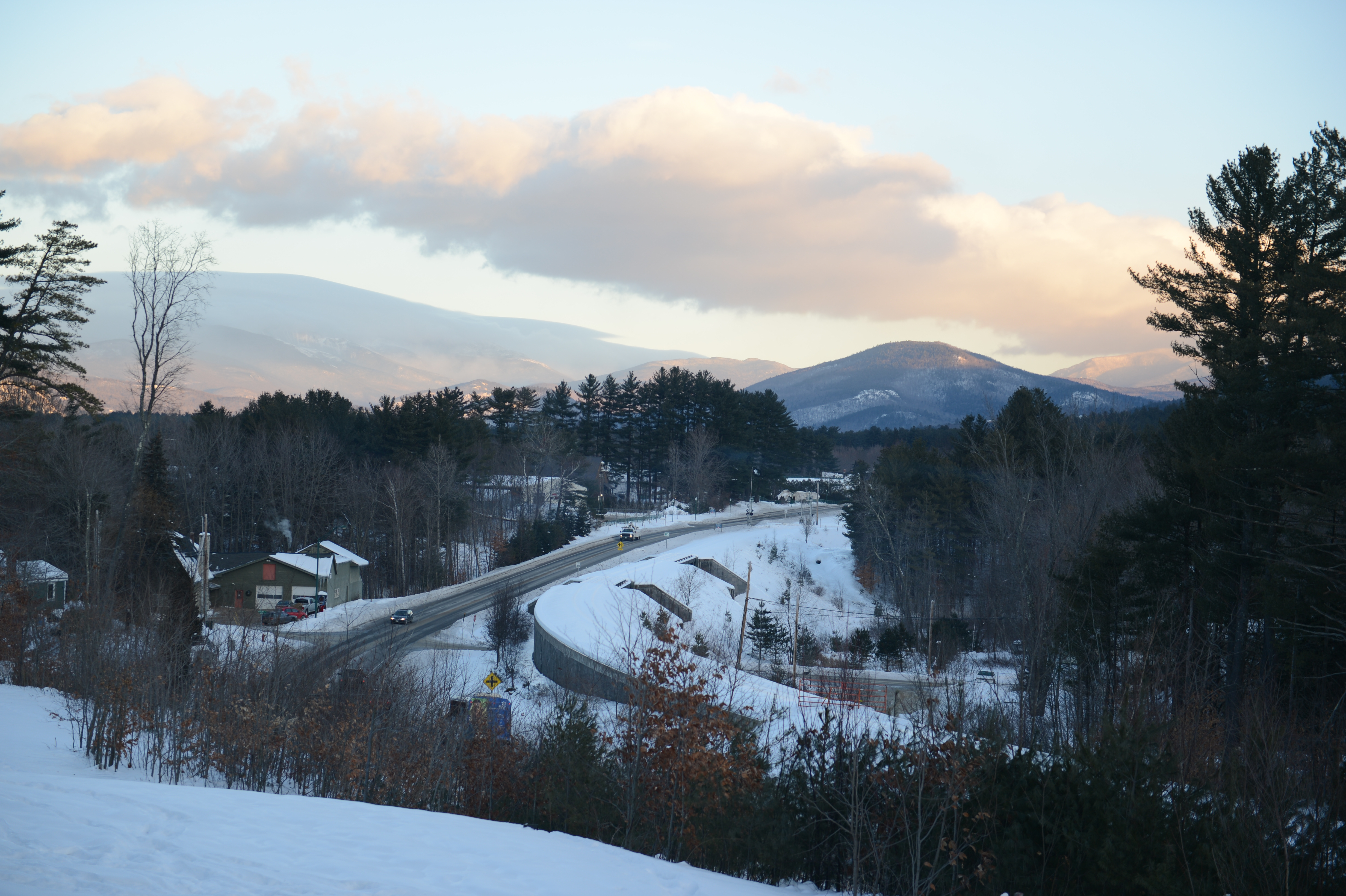 A snowy road curves through a winter landscape with mountains in the background and cars driving along.