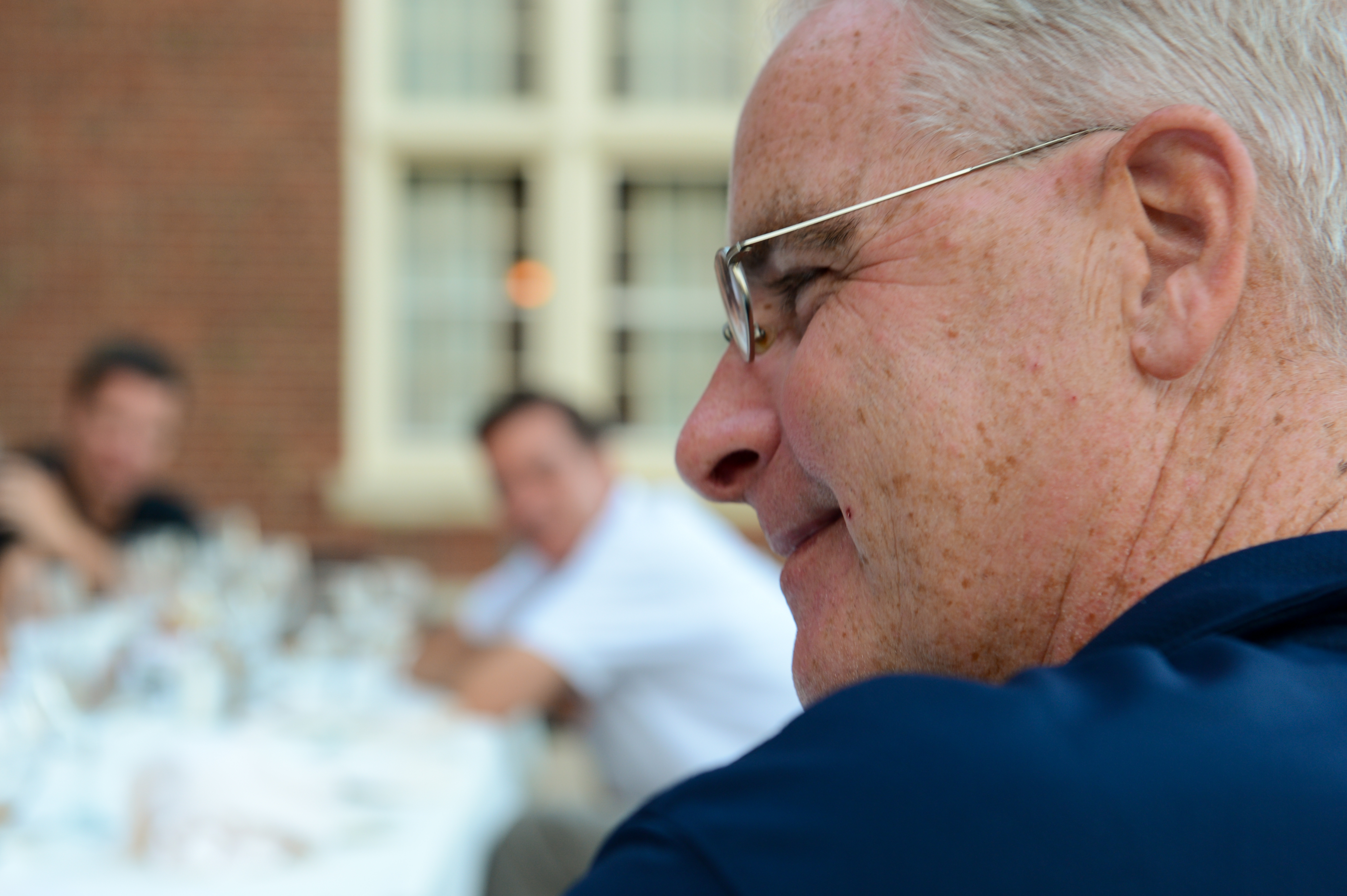 A man wearing glasses and a navy shirt smiles while seated at an outdoor dinner with others in the background.