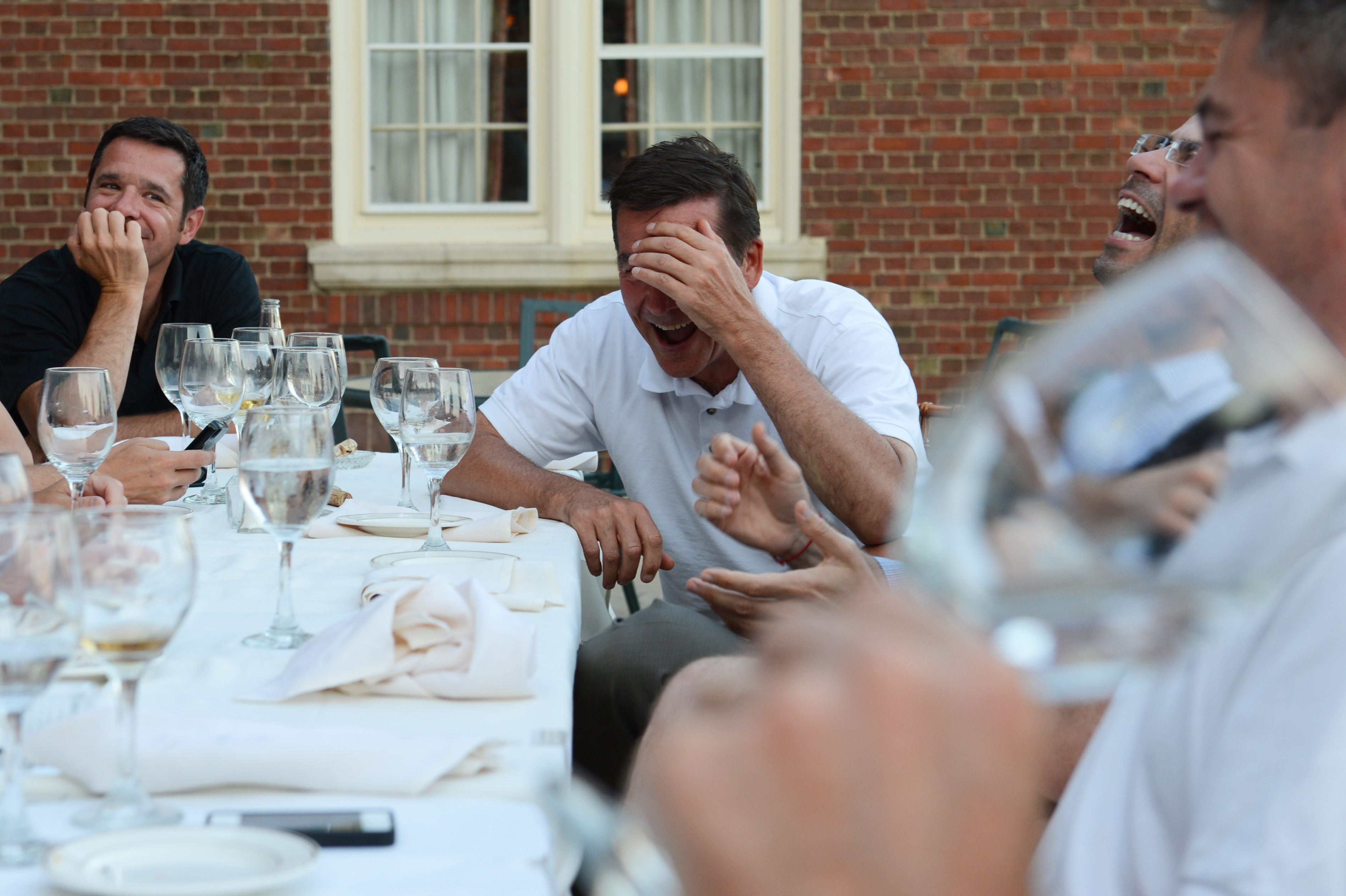 A group of people at an outdoor dinner table laughing, with one person covering their face while laughing.