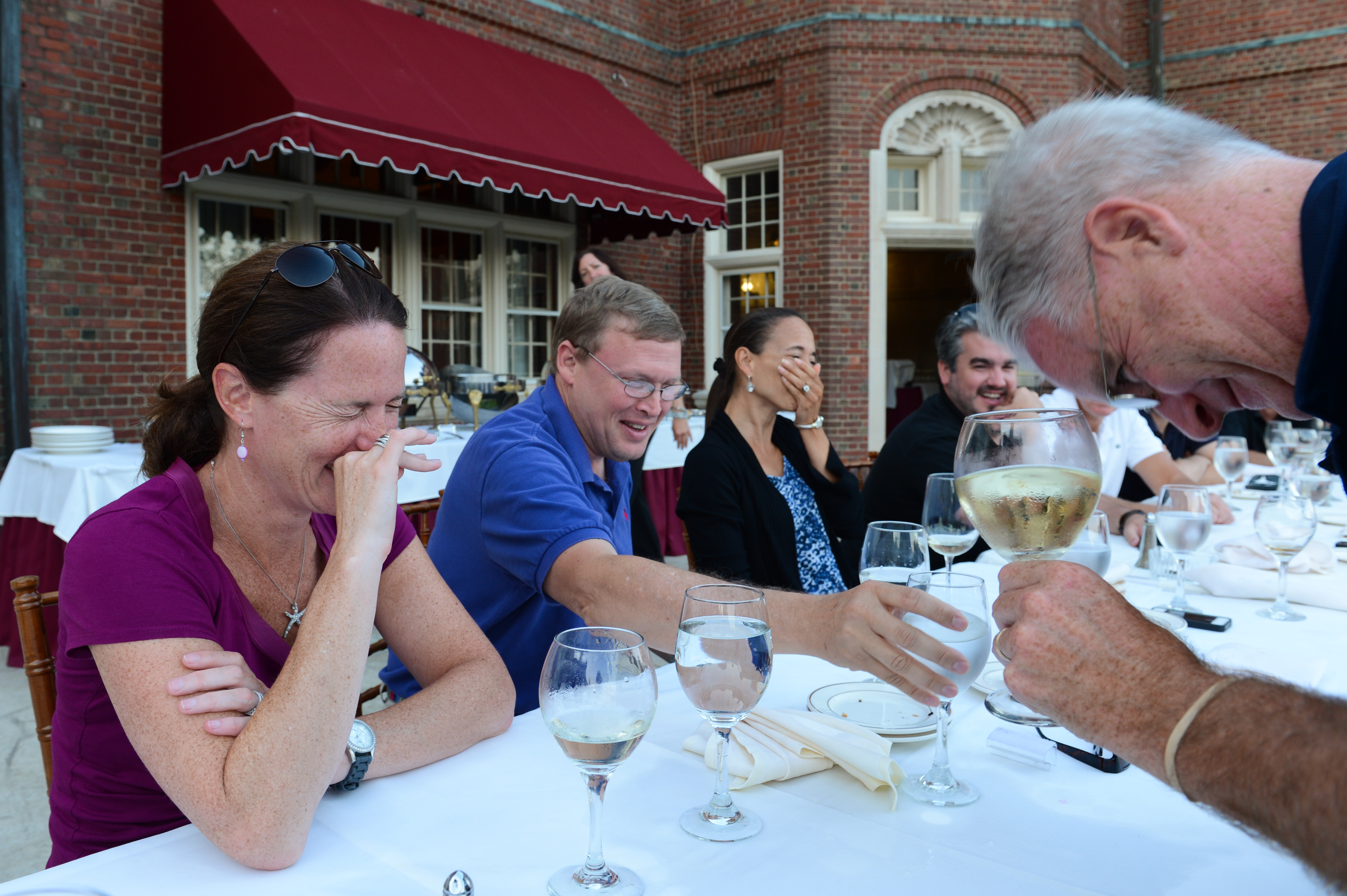 A group of people at an outdoor dinner table laughing, with one person leaning forward holding a wine glass.