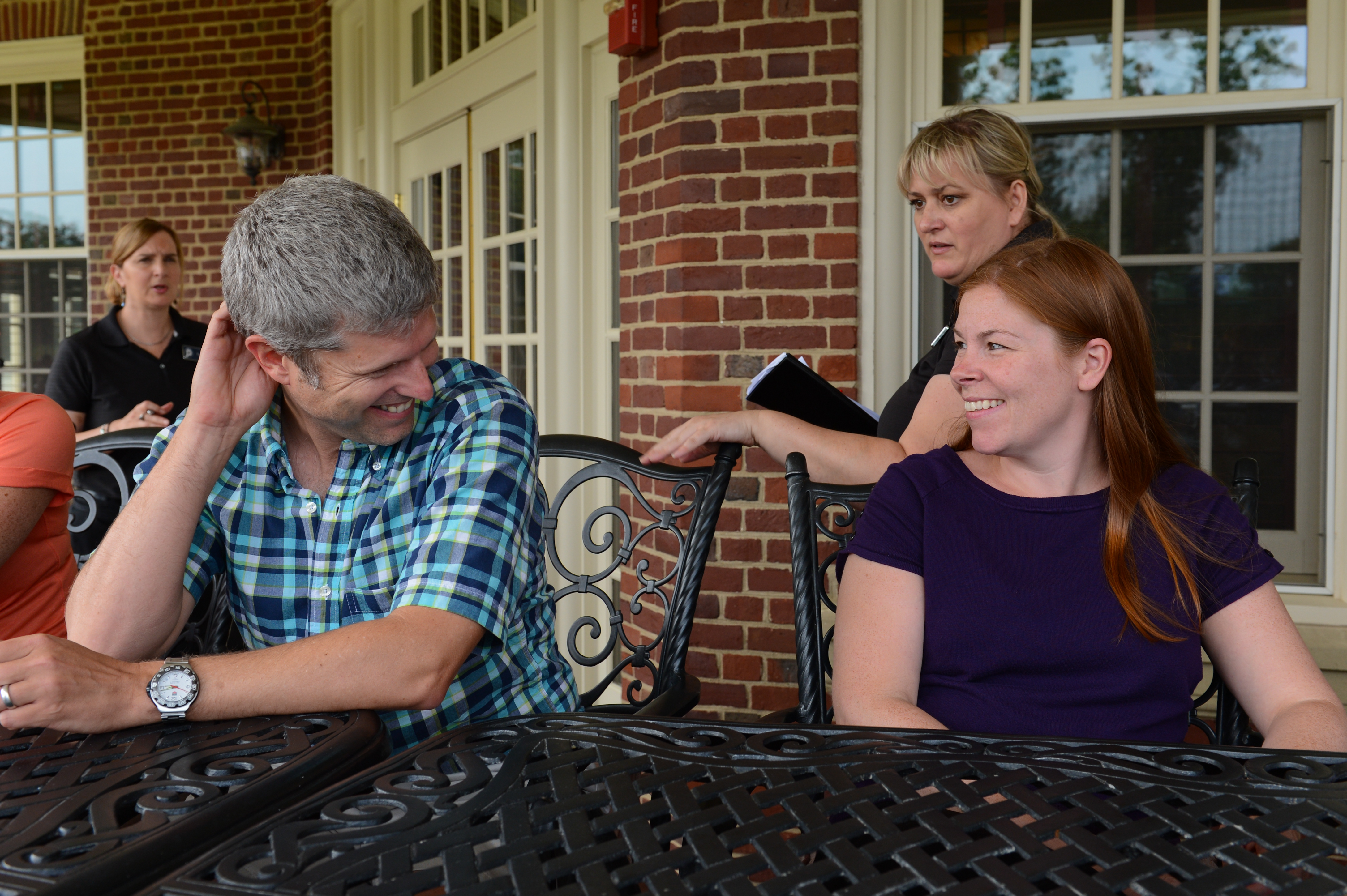 A man and a woman sit at an outdoor table, smiling at each other, while two others stand nearby.