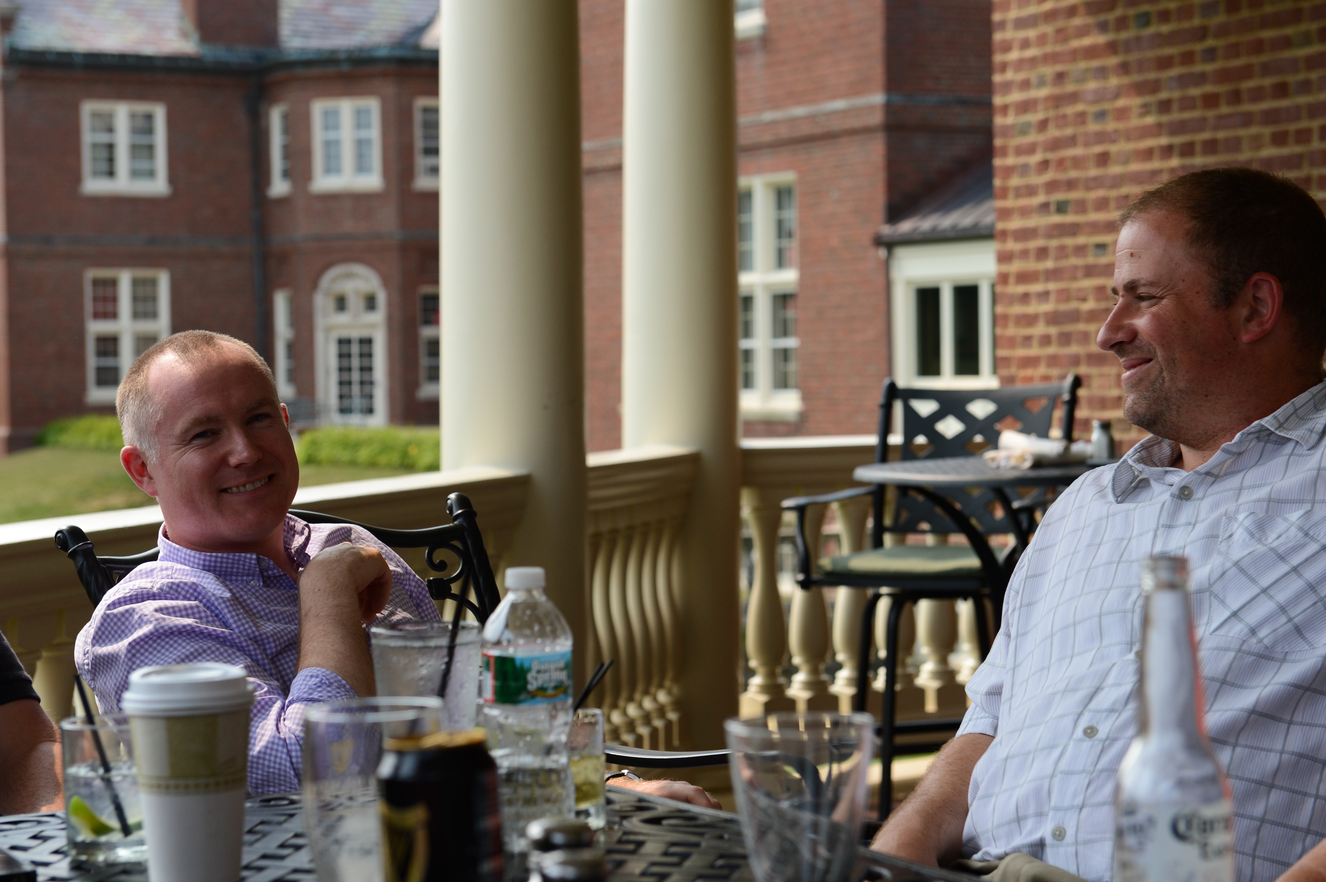 Two men sit on a patio, smiling and chatting, with various drinks on the table in front of them.