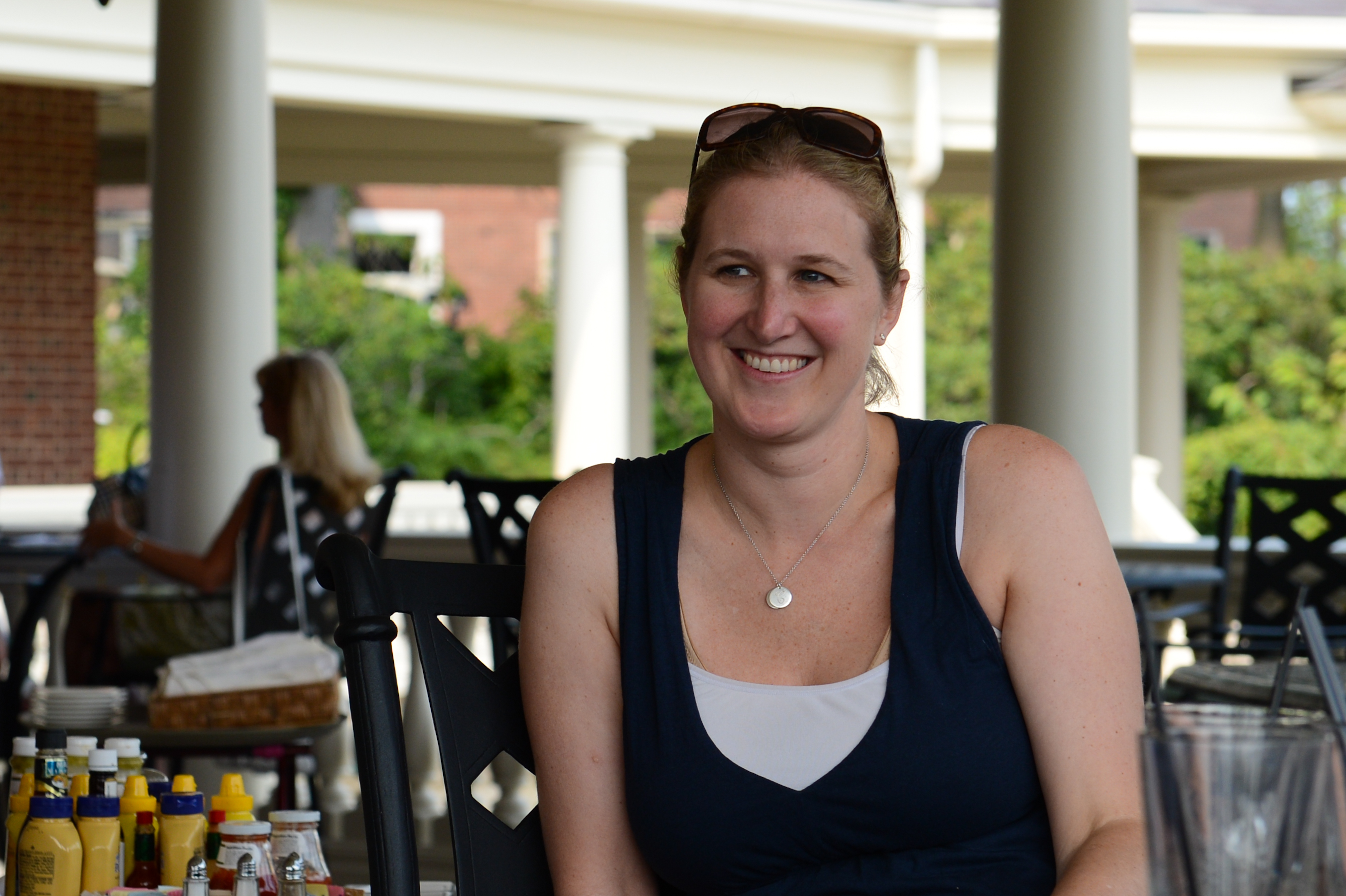A woman with sunglasses on her head smiles while sitting at an outdoor table with drinks and condiments.