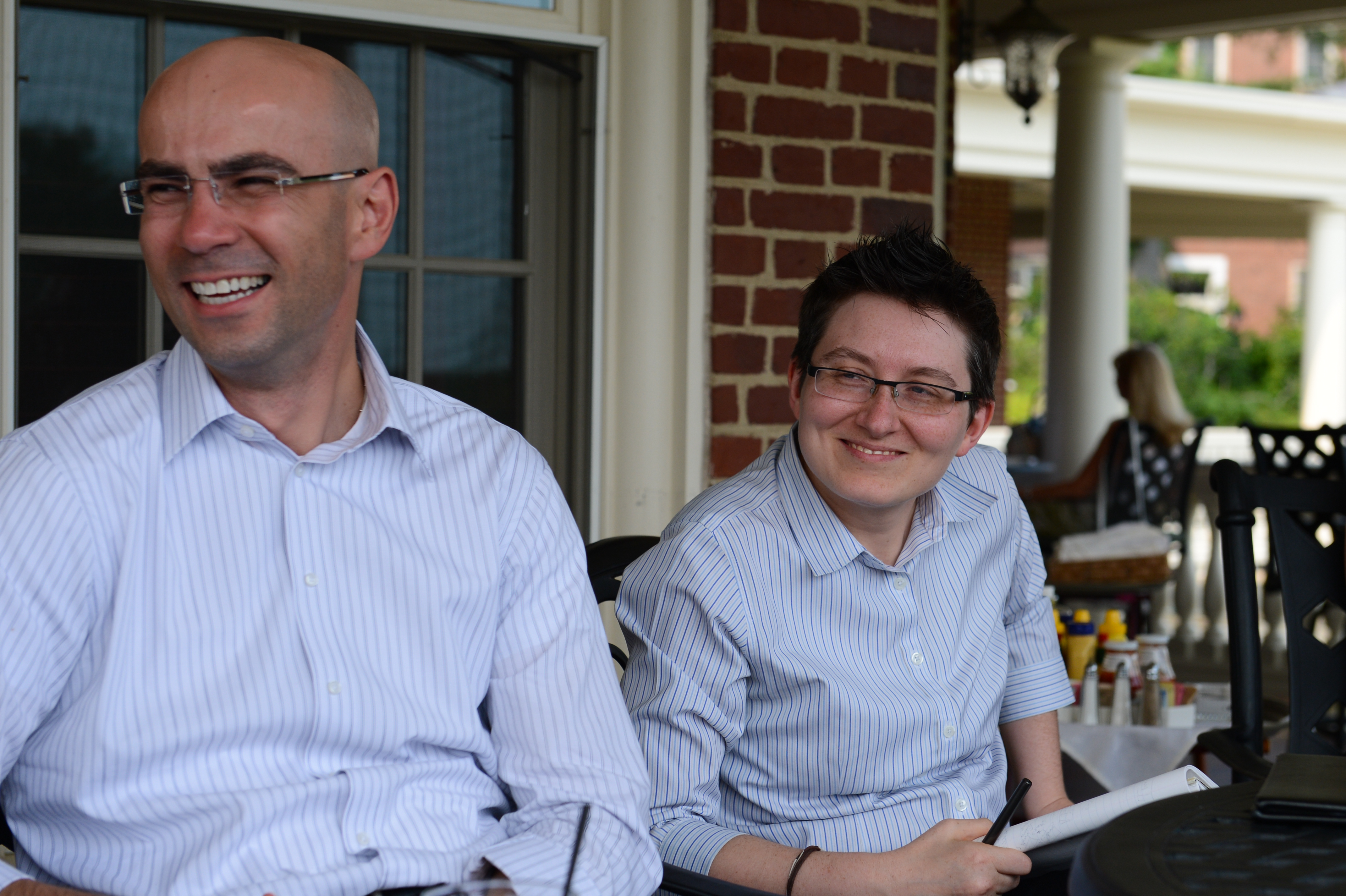 Two people in striped shirts are sitting outside, smiling and chatting while holding drinks and a notebook.