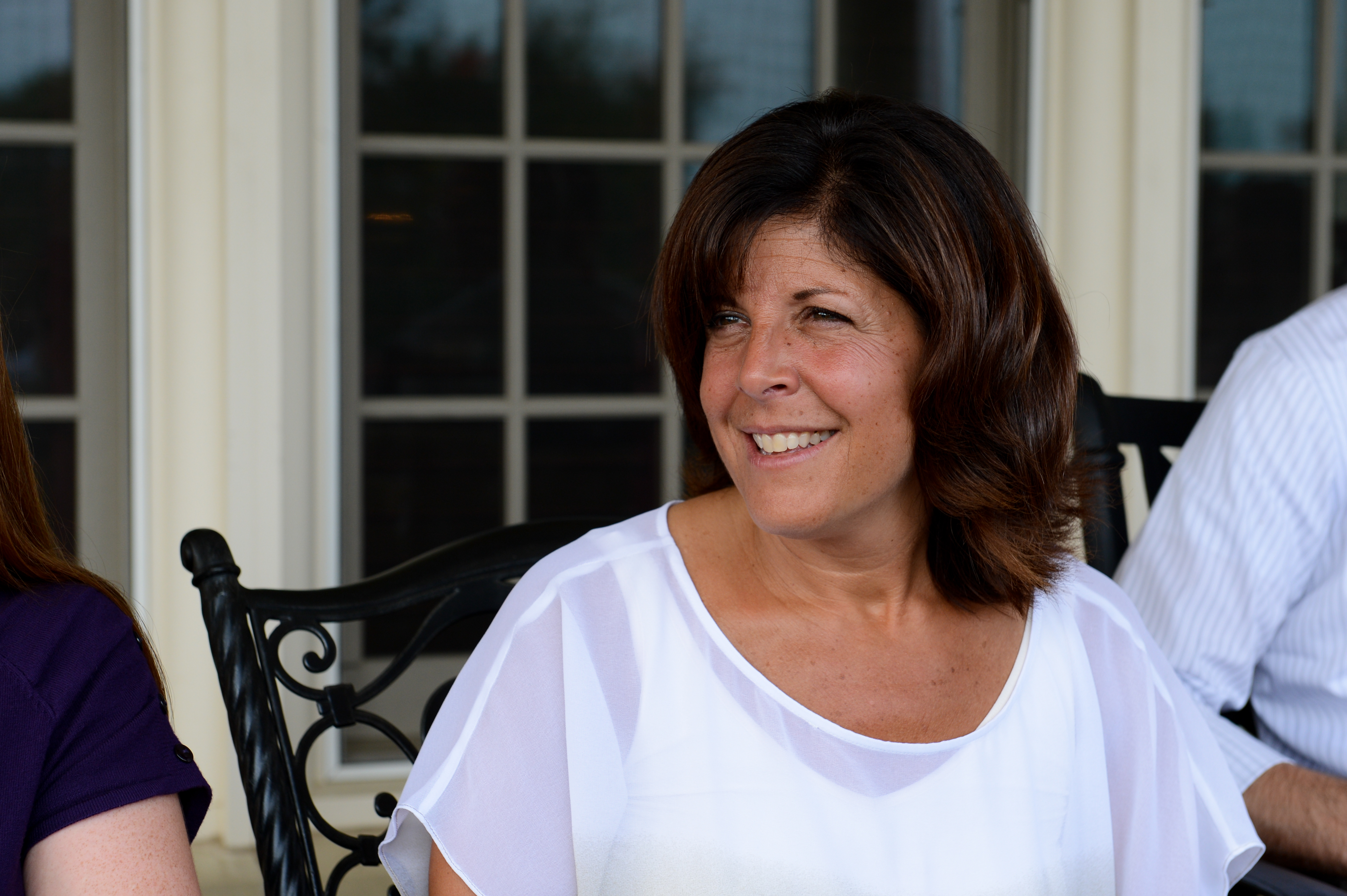 A woman in a white blouse smiles while sitting outdoors with others at a gathering.