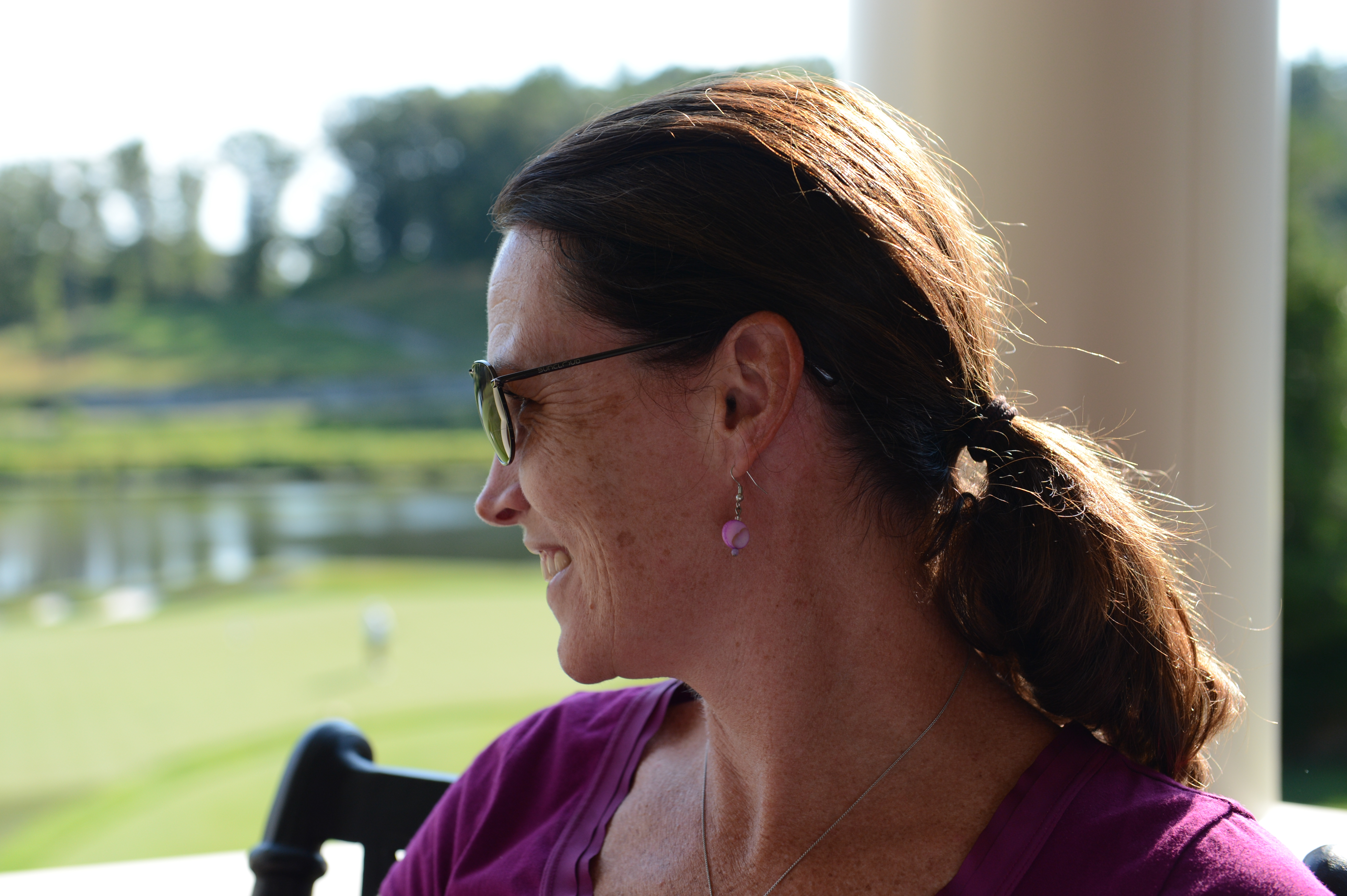 A woman wearing sunglasses and a purple top sits outdoors, looking to the side with a relaxed expression.
