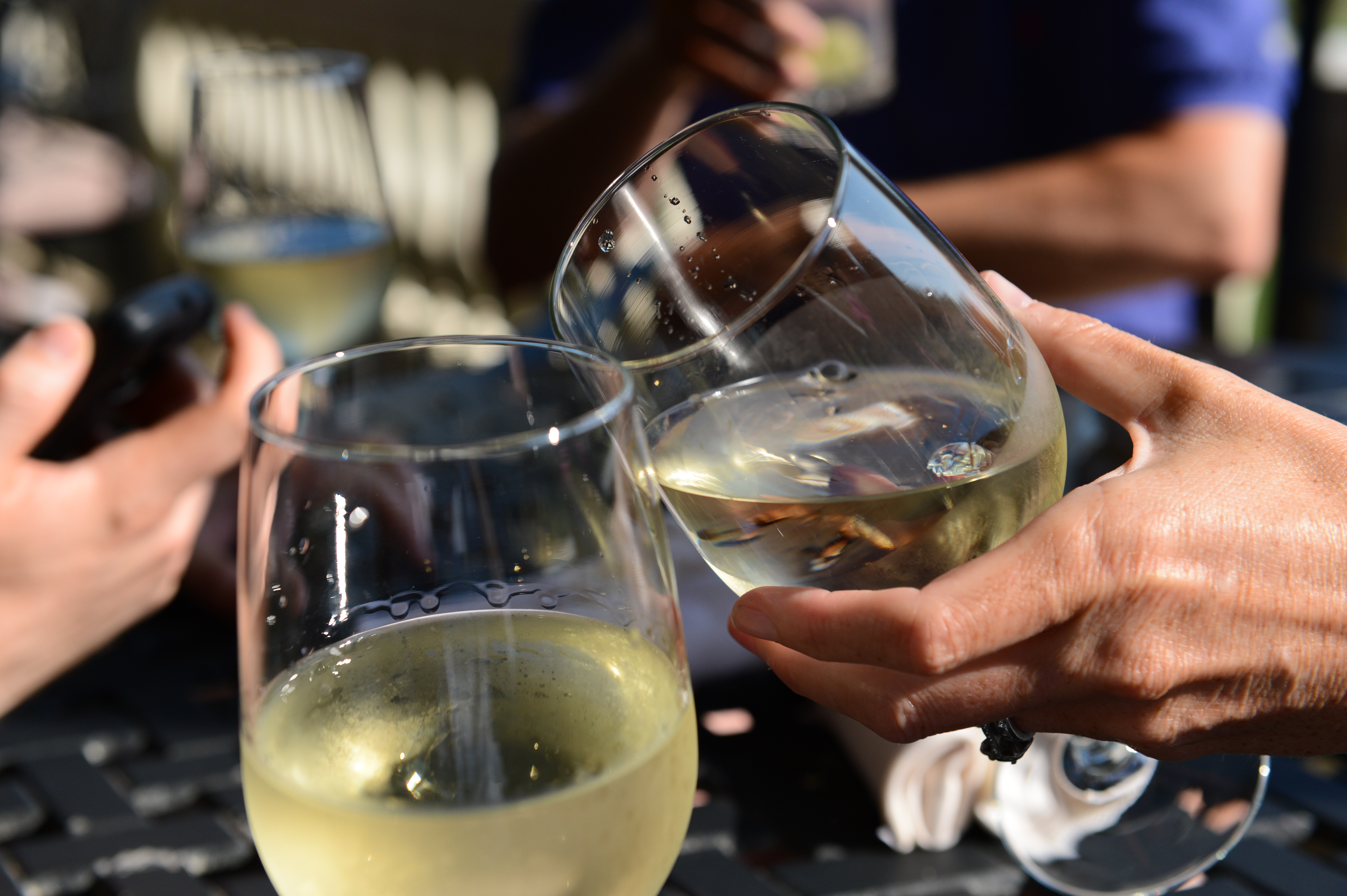 Two people clink glasses of white wine together while others hold drinks in the background.