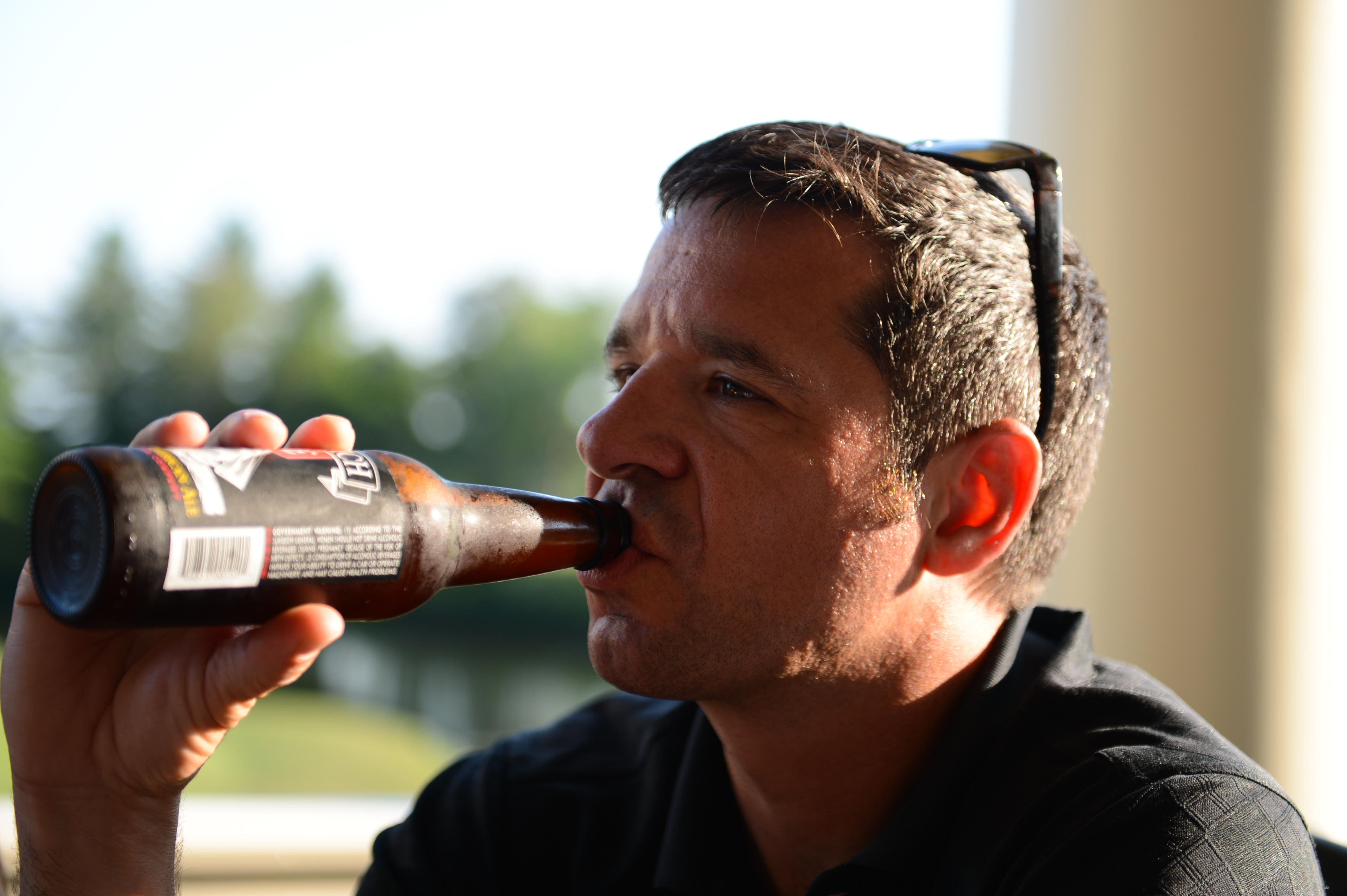 A man in a black shirt drinks from a brown beer bottle while sitting outdoors.