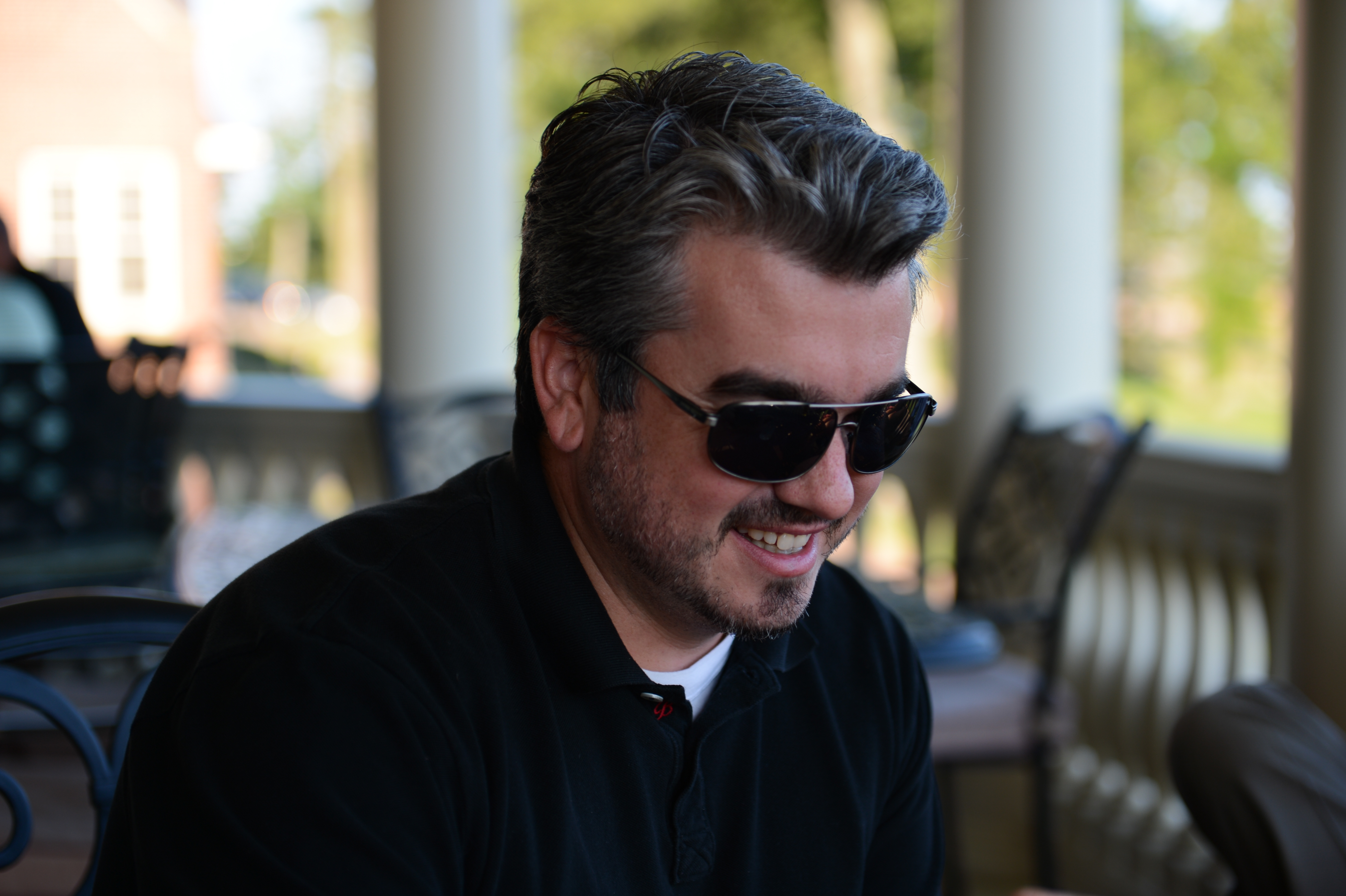 A man wearing sunglasses and a black shirt smiles while sitting outdoors at a gathering.