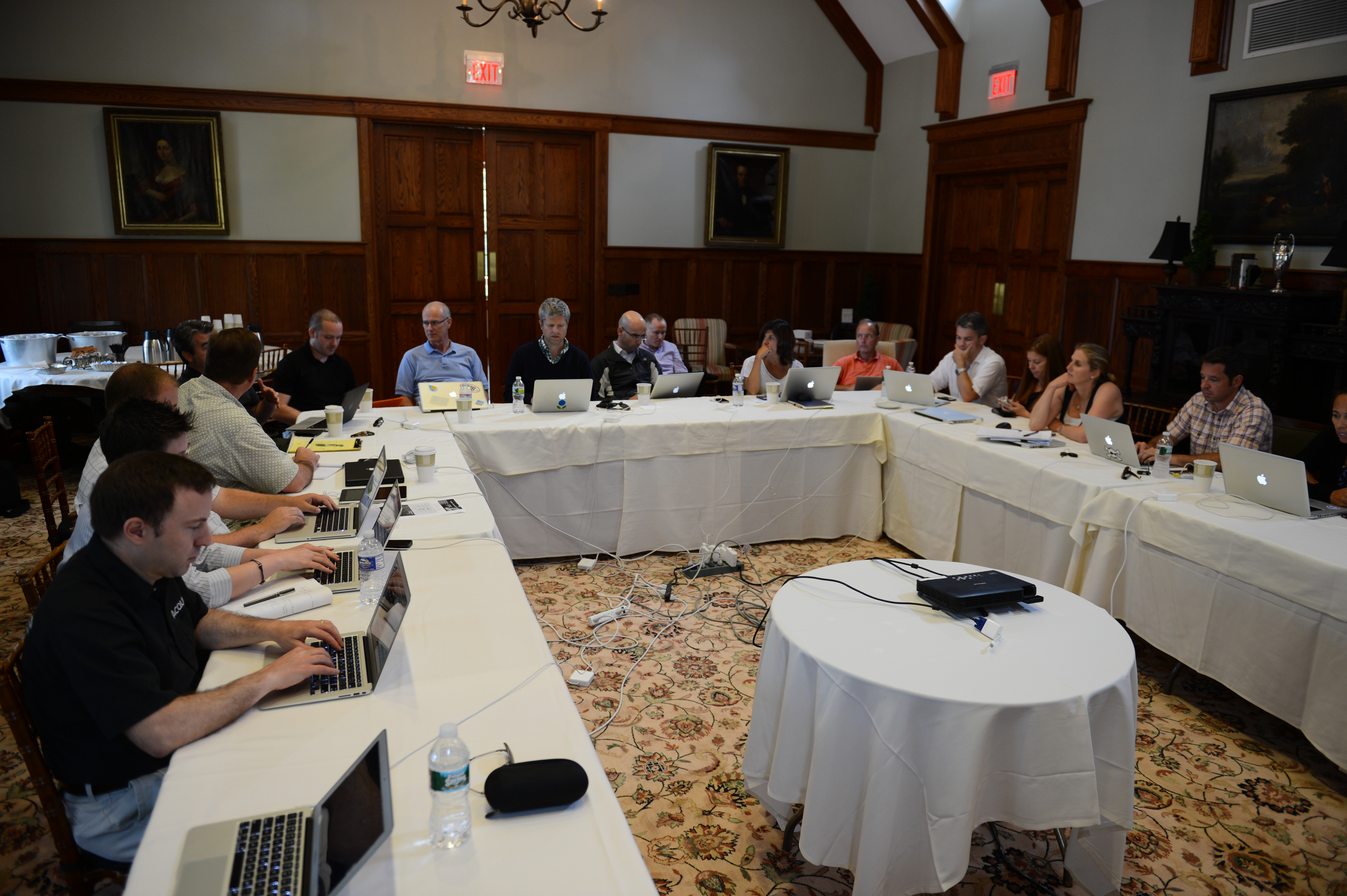 A group of people sitting around a U-shaped table in a meeting, using laptops and taking notes.