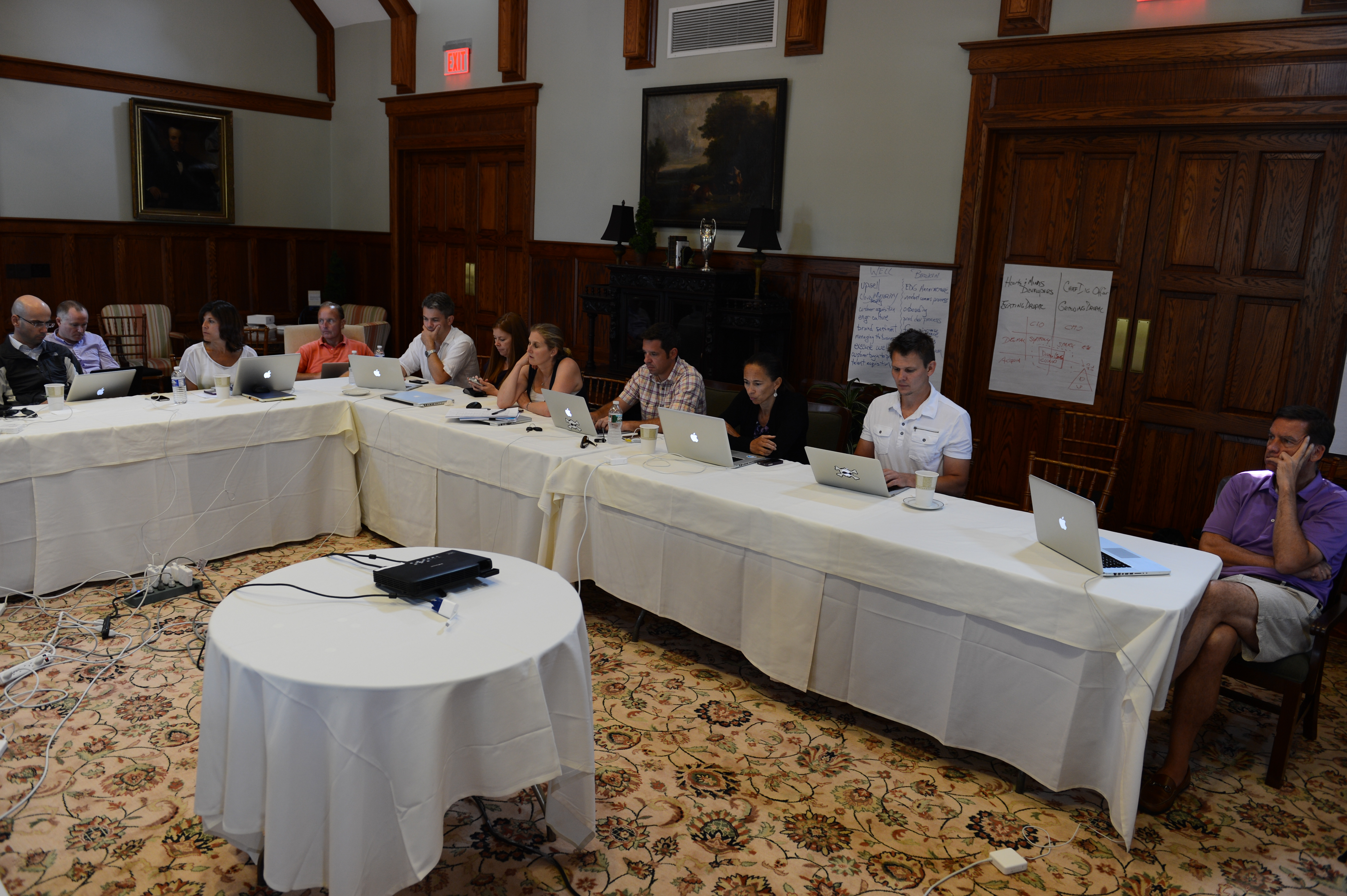 A group of people sit around tables with laptops, engaged in a meeting and discussion.