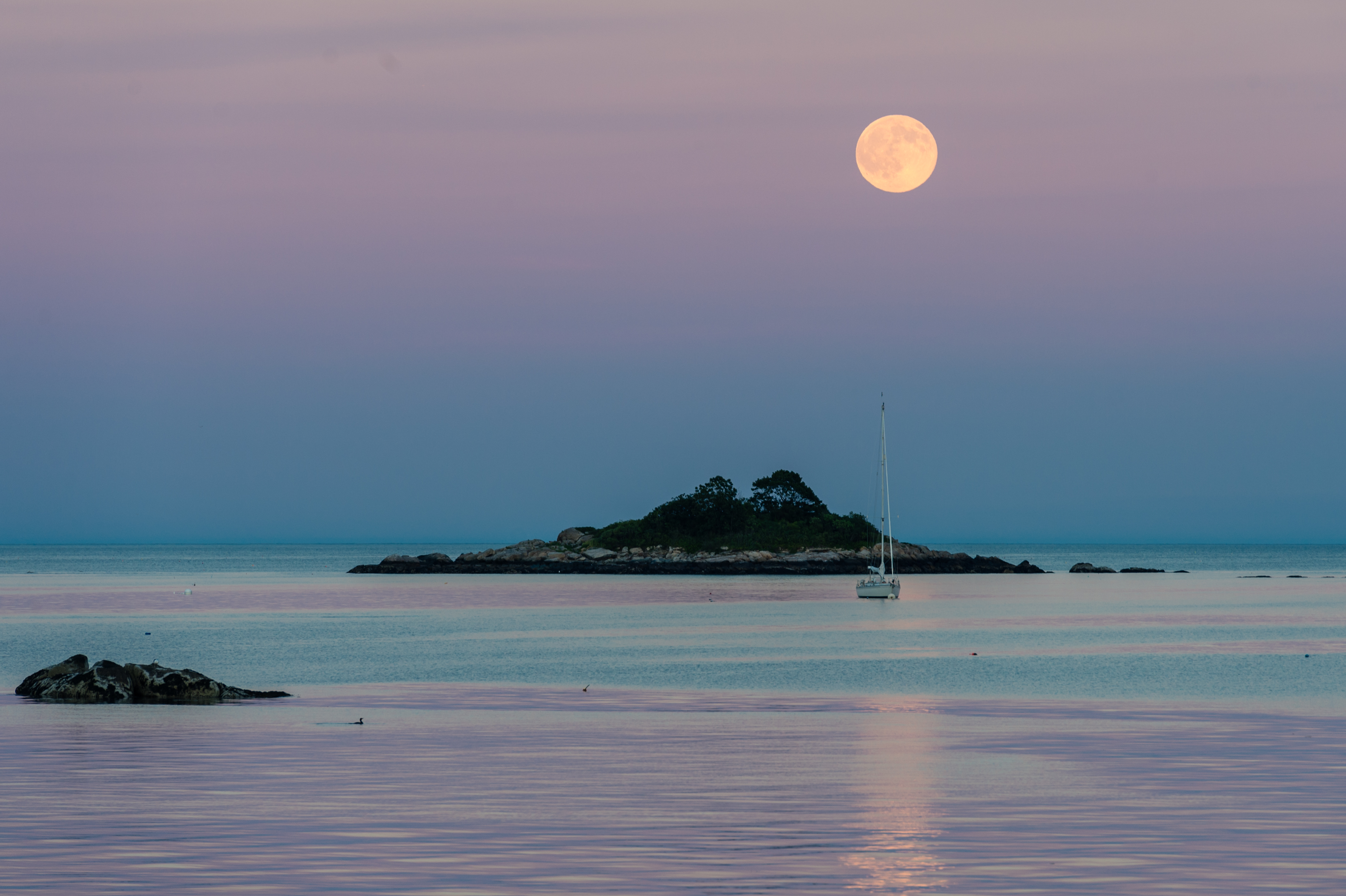 A full moon shines over calm ocean waters, with a small island and a sailboat in the distance.
