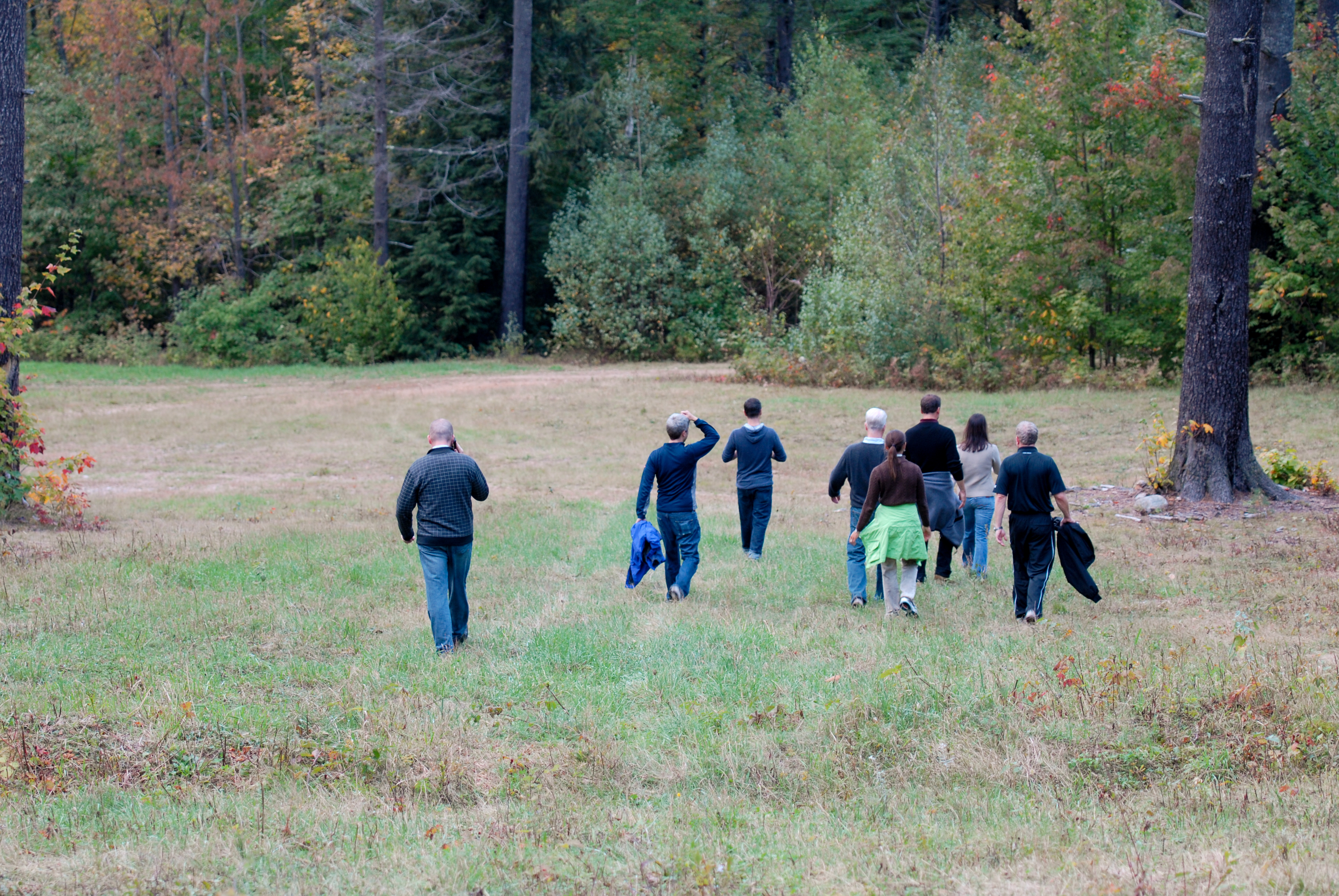 A group of people walks through a grassy field toward a wooded area, some carrying jackets.