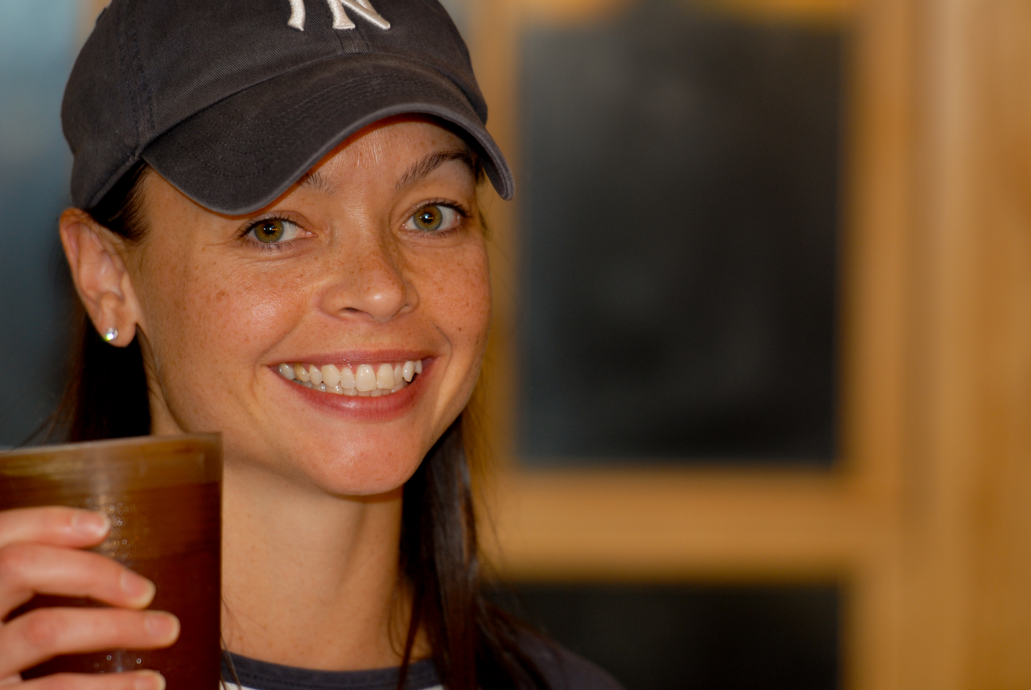 A smiling person wearing a baseball cap holds up a drink in a casual indoor setting.
