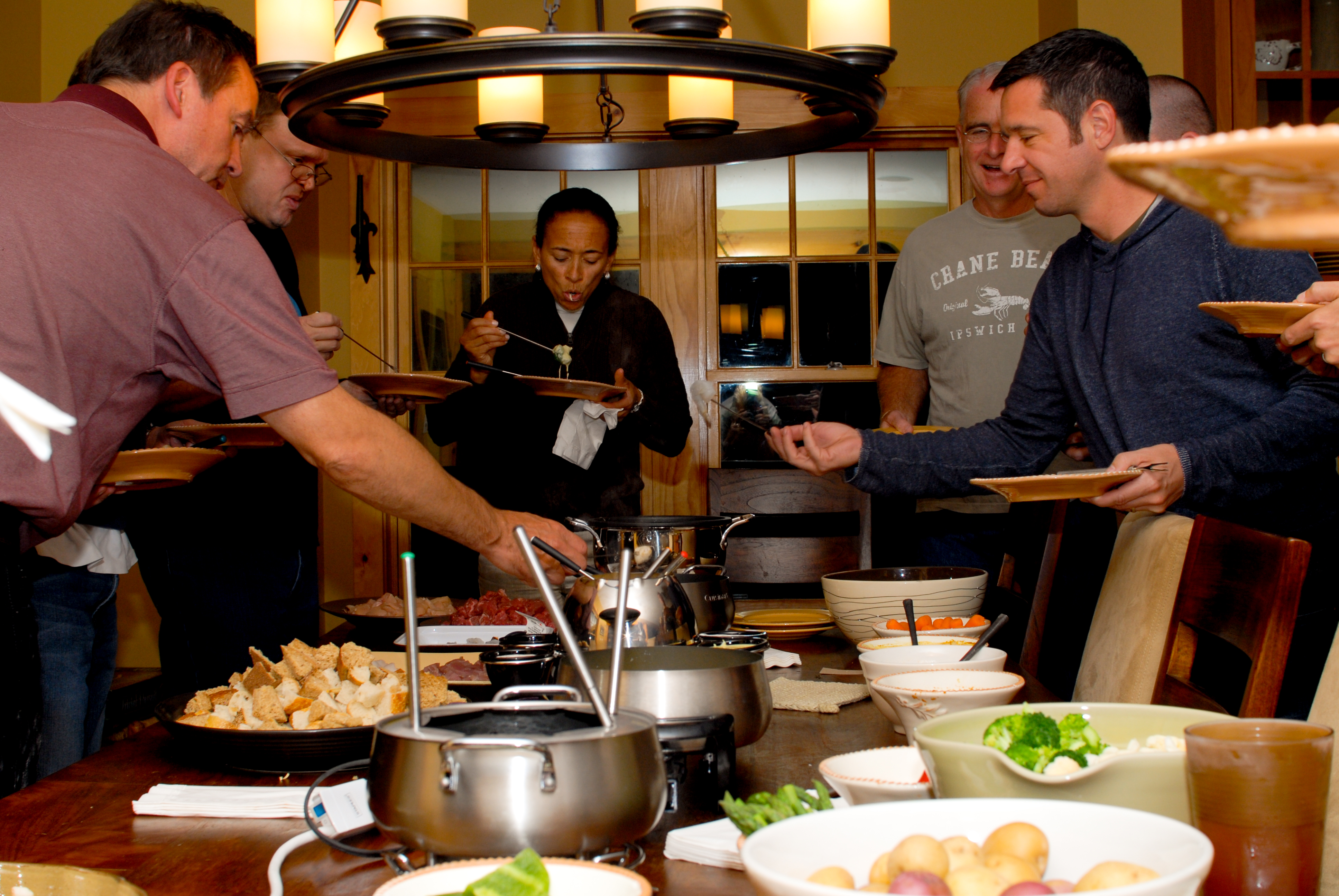 A group of people gathers around a table, serving themselves food from a fondue spread.