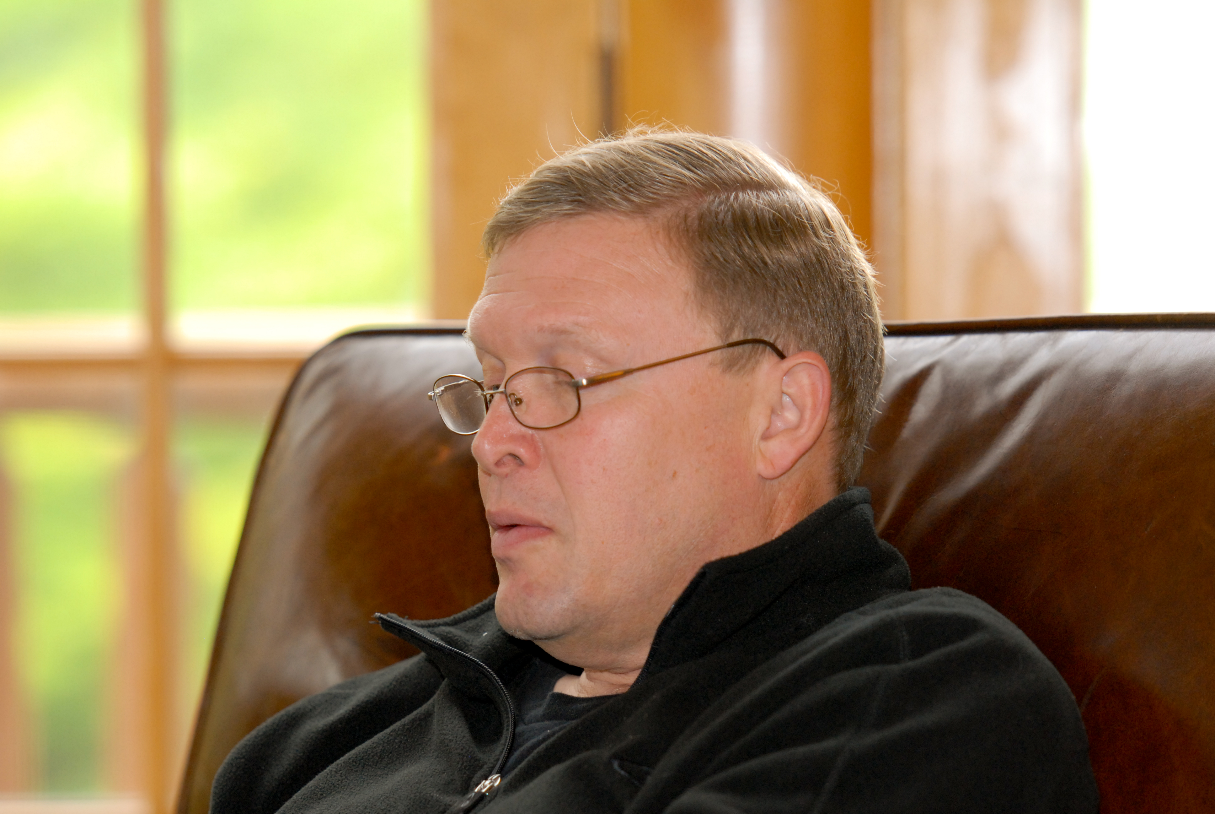A man wearing glasses and a black jacket sits on a leather chair, looking down with a focused expression.