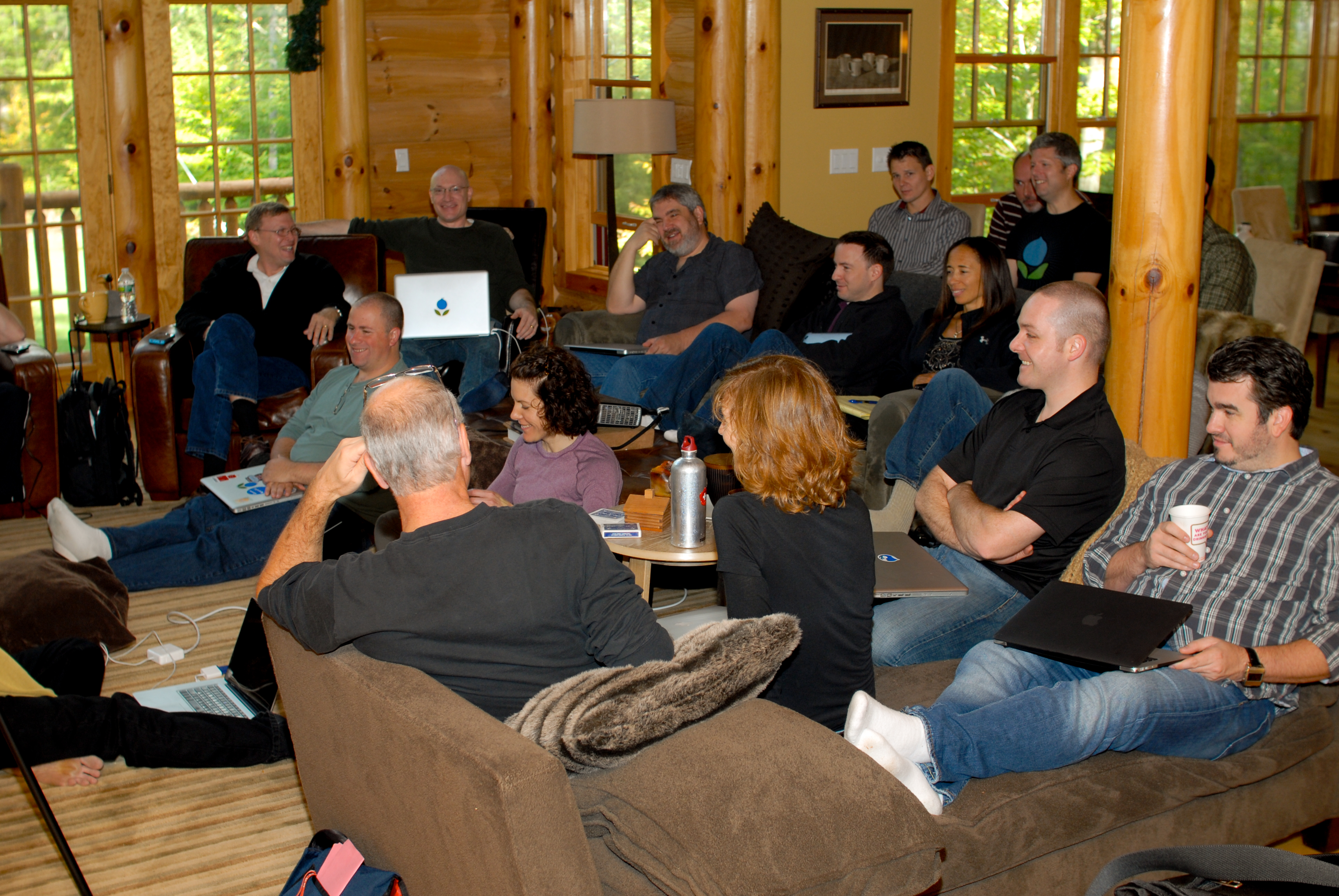 A group of people in a cozy cabin setting, sitting with laptops and drinks, engaged in a discussion.