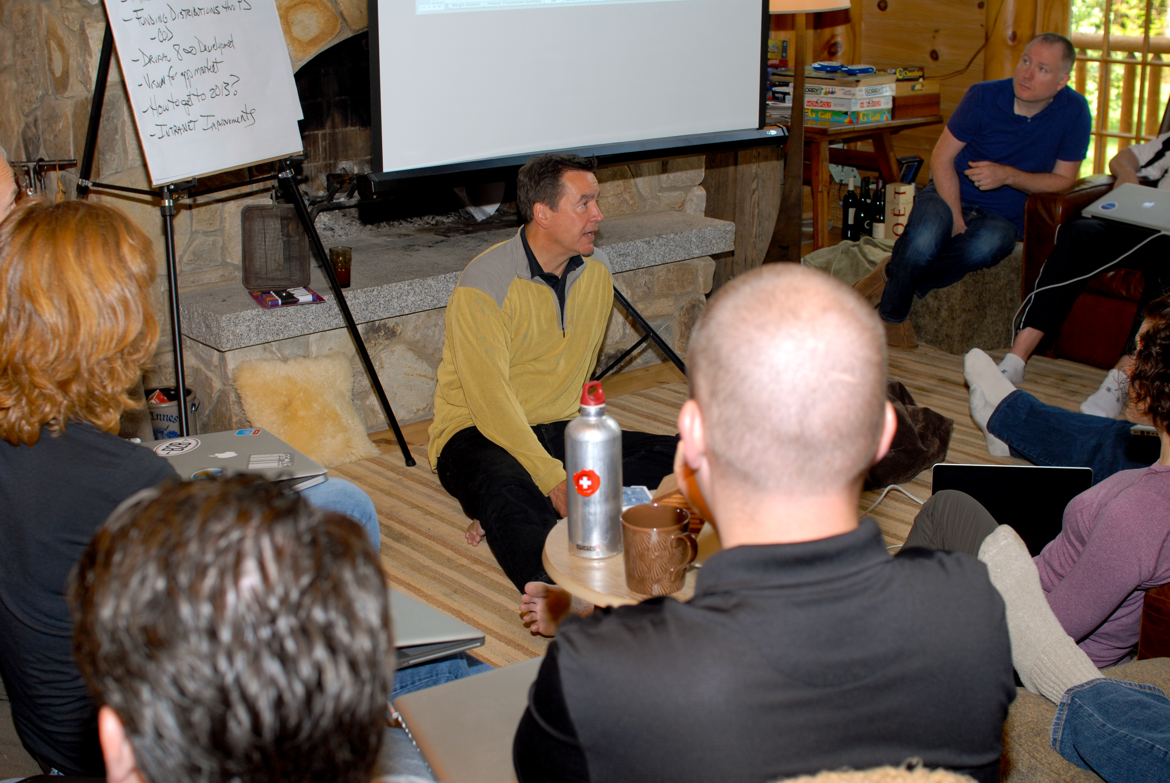 A man sits on the floor leading a discussion while a group of people listens in a casual setting.
