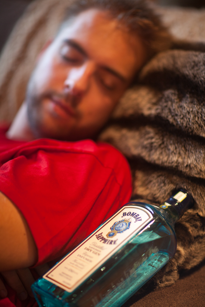 A person in a red shirt is sleeping on a couch next to a bottle of Bombay Sapphire gin.