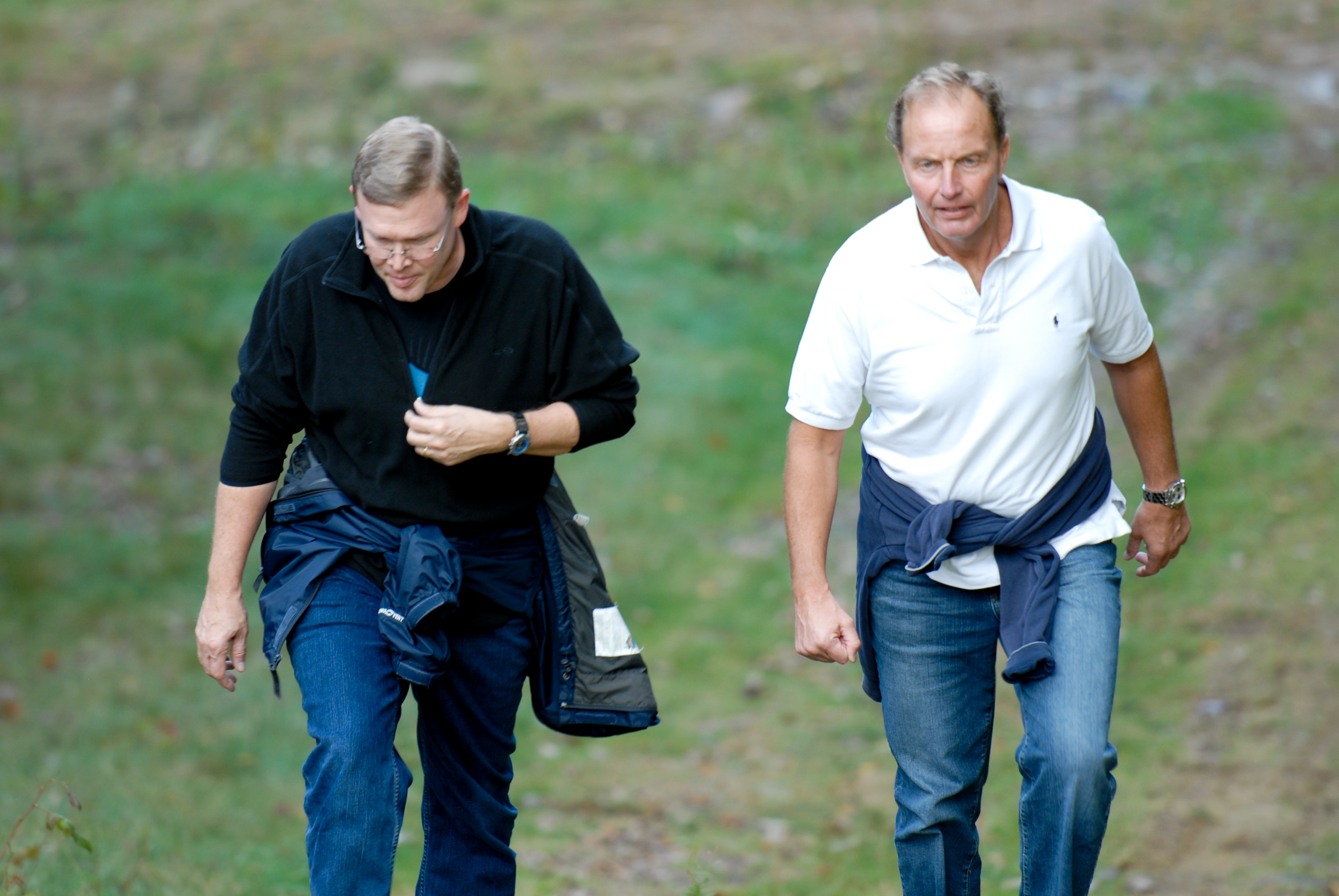 Two men walk uphill on a grassy path, both wearing jackets tied around their waists.