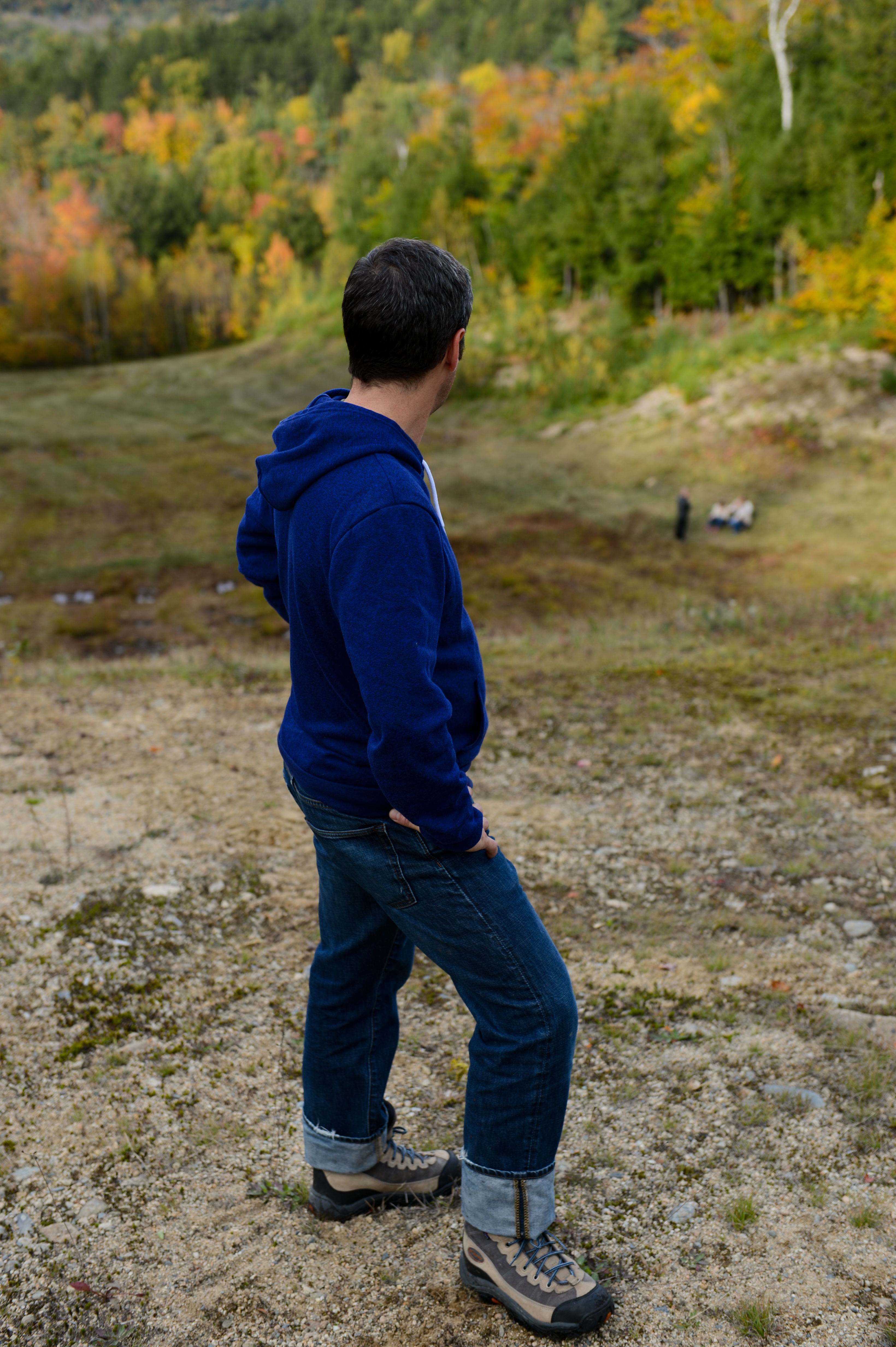 A person in a blue hoodie stands on a hillside, looking down at a group in the distance.