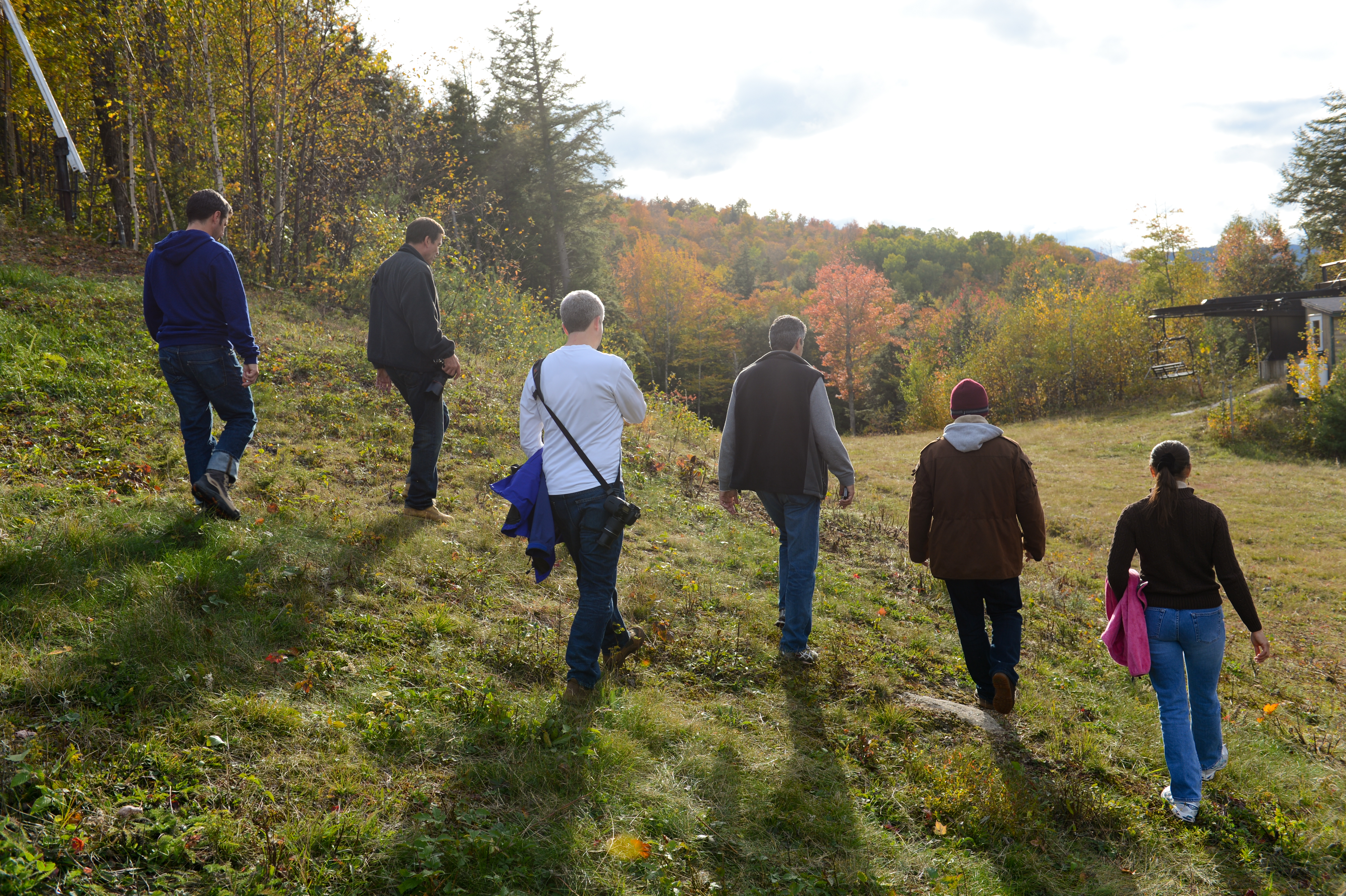 A group of six people walks uphill on a grassy path surrounded by autumn trees.