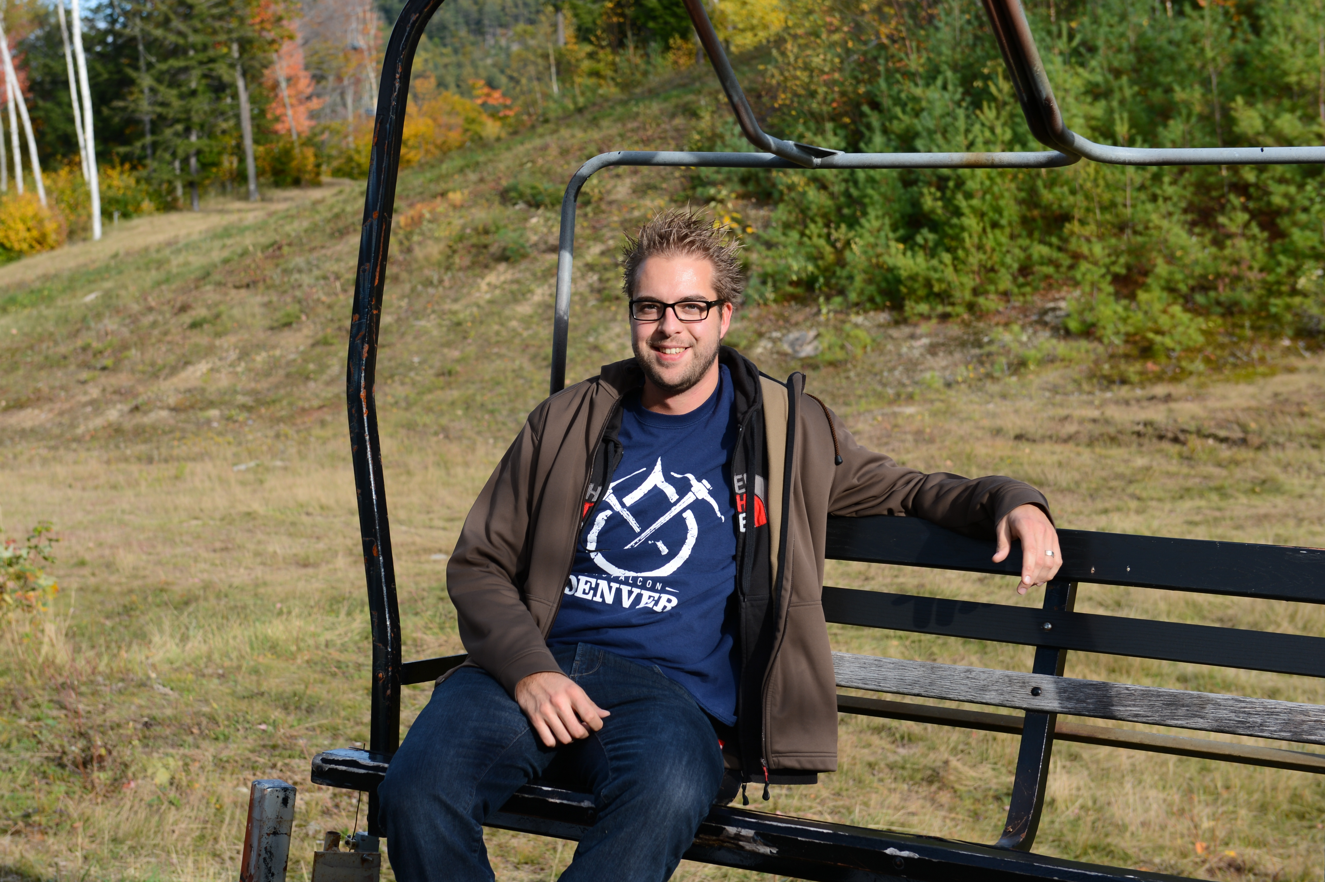 A man wearing glasses and a jacket sits on a ski lift bench, smiling at the camera.