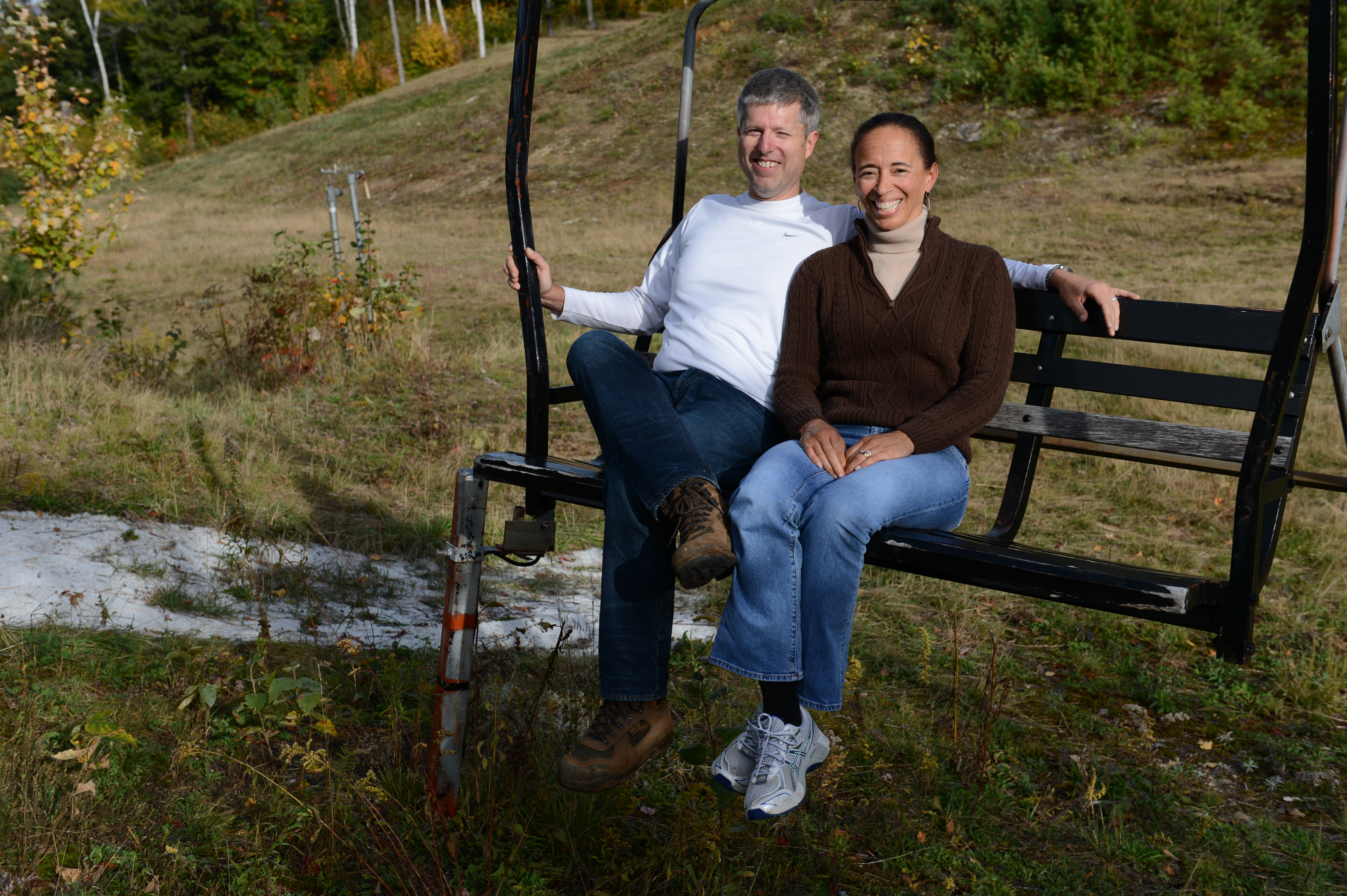 Two people sit on a stationary ski lift chair outdoors, smiling at the camera.