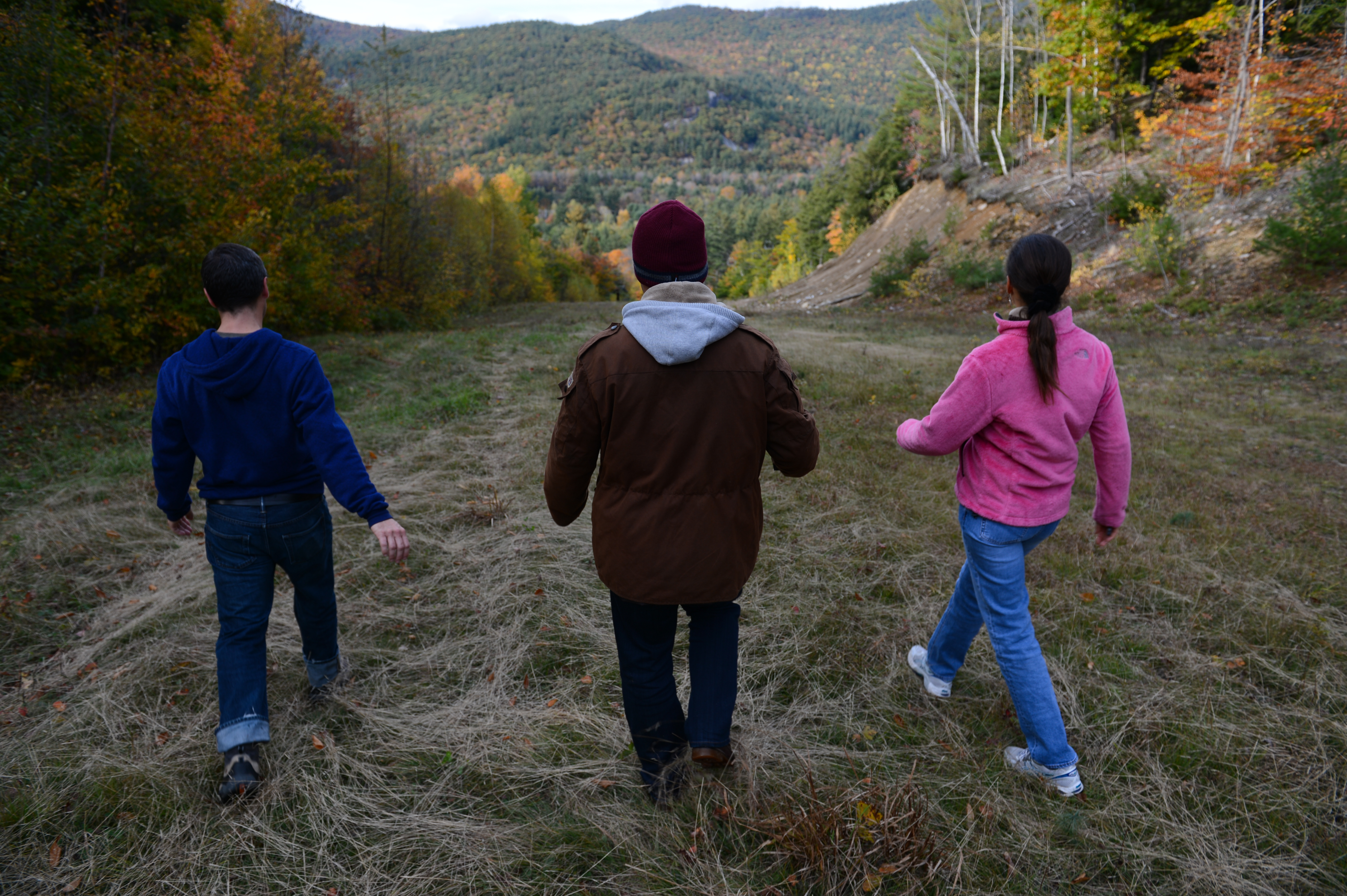 Three people walk through a grassy hillside surrounded by trees with autumn colors.
