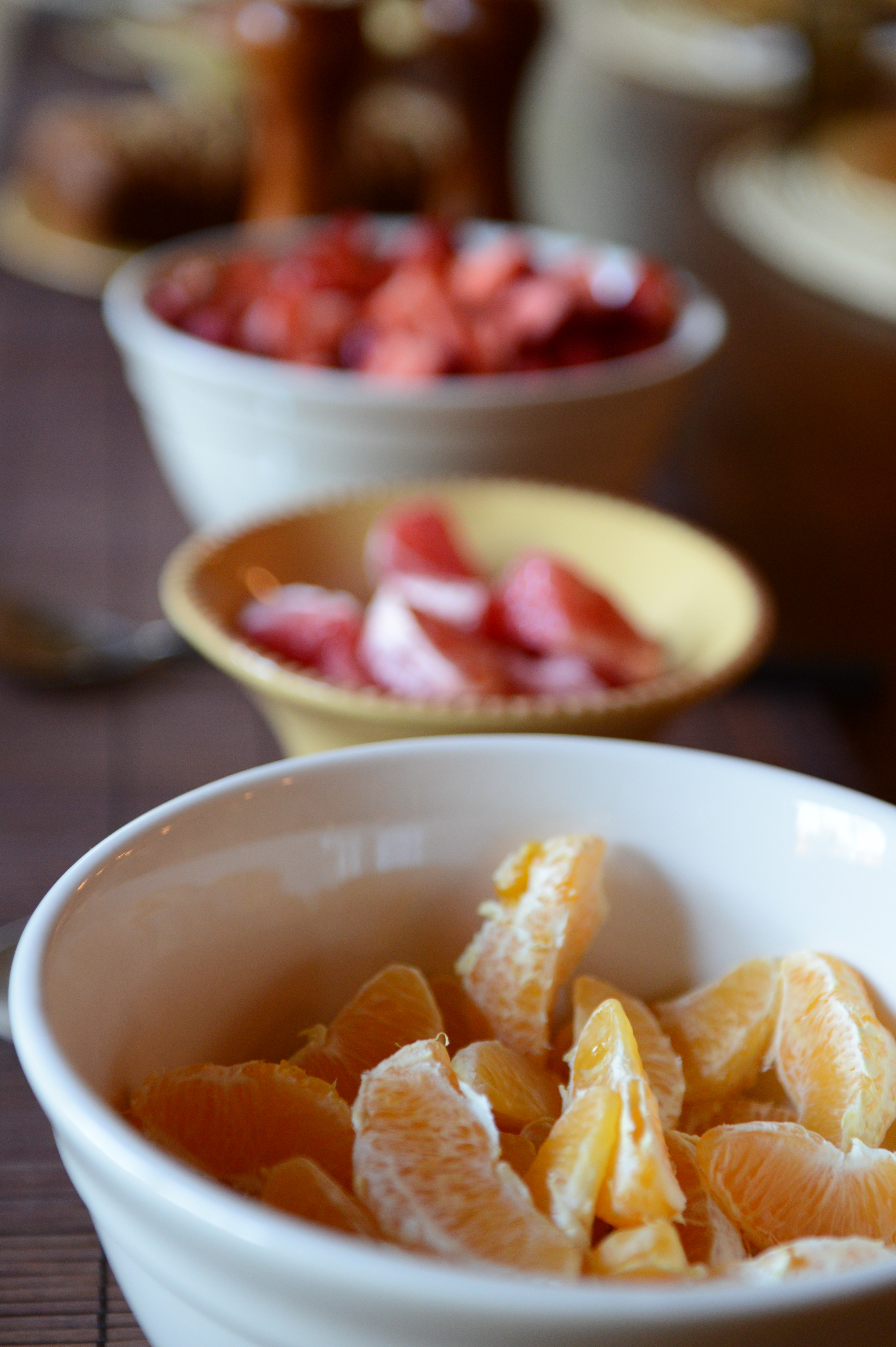 A white bowl filled with peeled orange slices sits in the foreground, with other fruit bowls in the background.