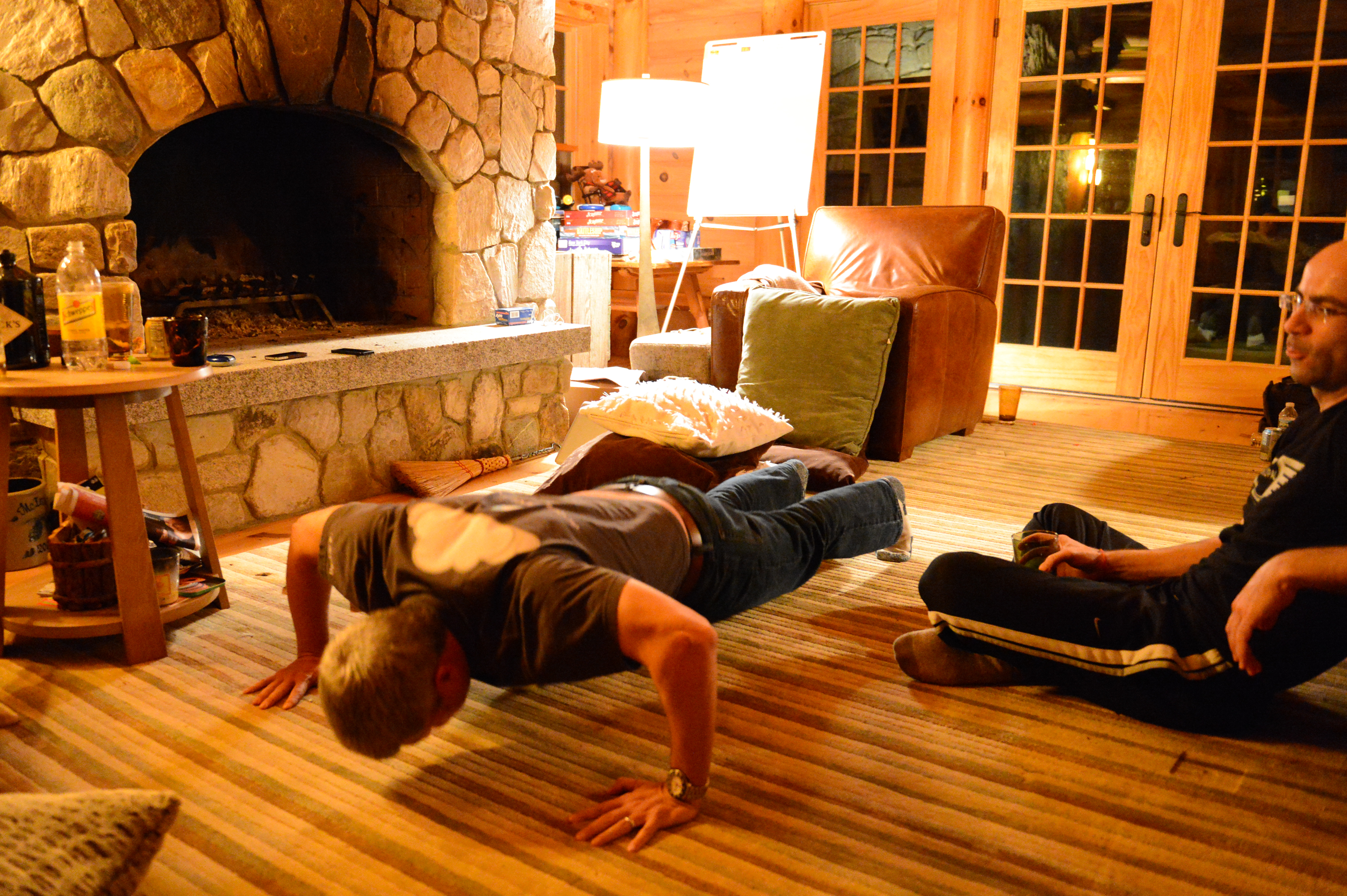 A man does push-ups on a rug while another person sits nearby watching in a cozy indoor setting.