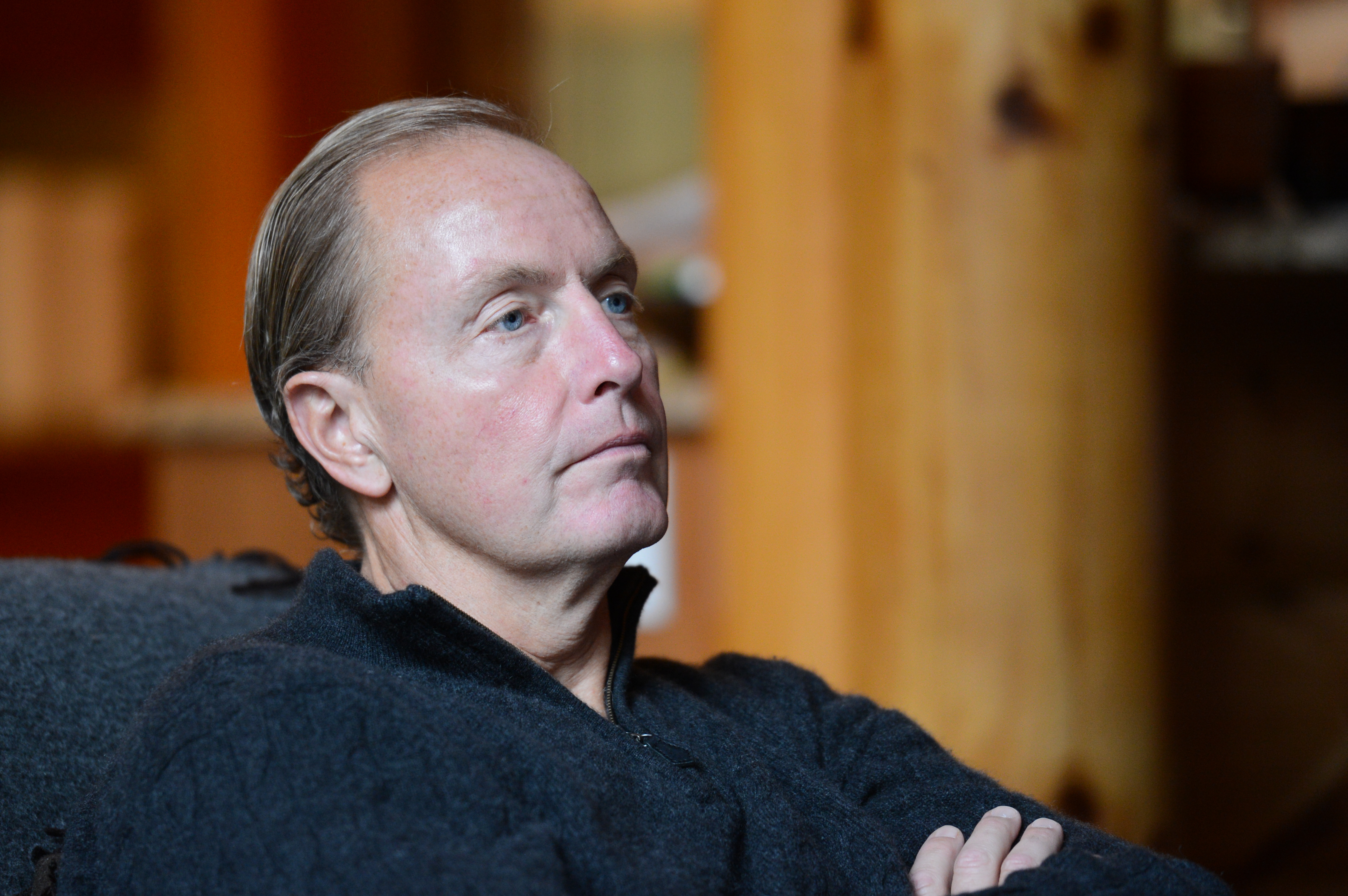 A man in a black sweater sits with arms crossed, attentively listening in a warmly lit indoor setting.