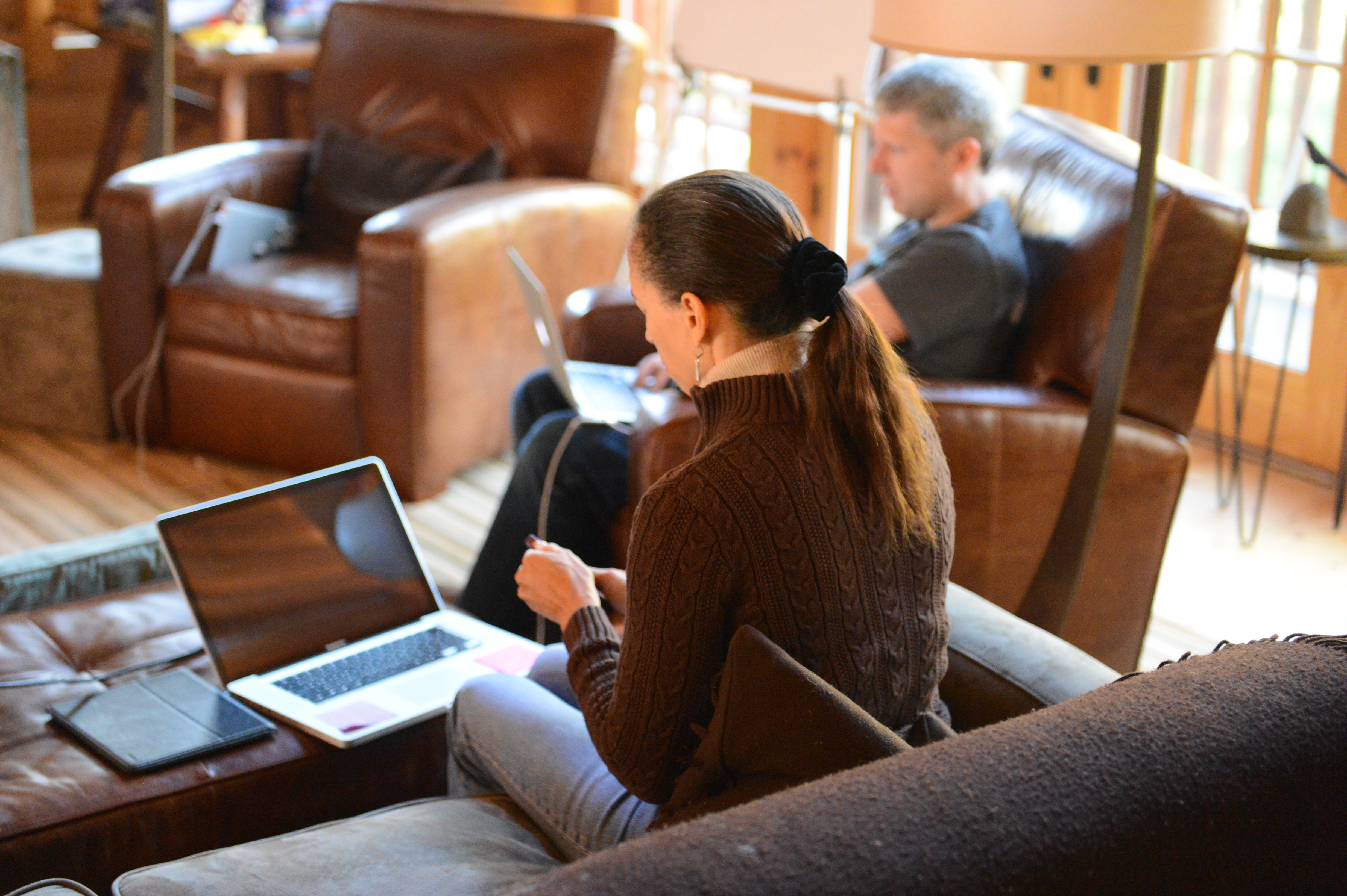 Two people working on laptops in a cozy lounge area with leather chairs and wooden surroundings.