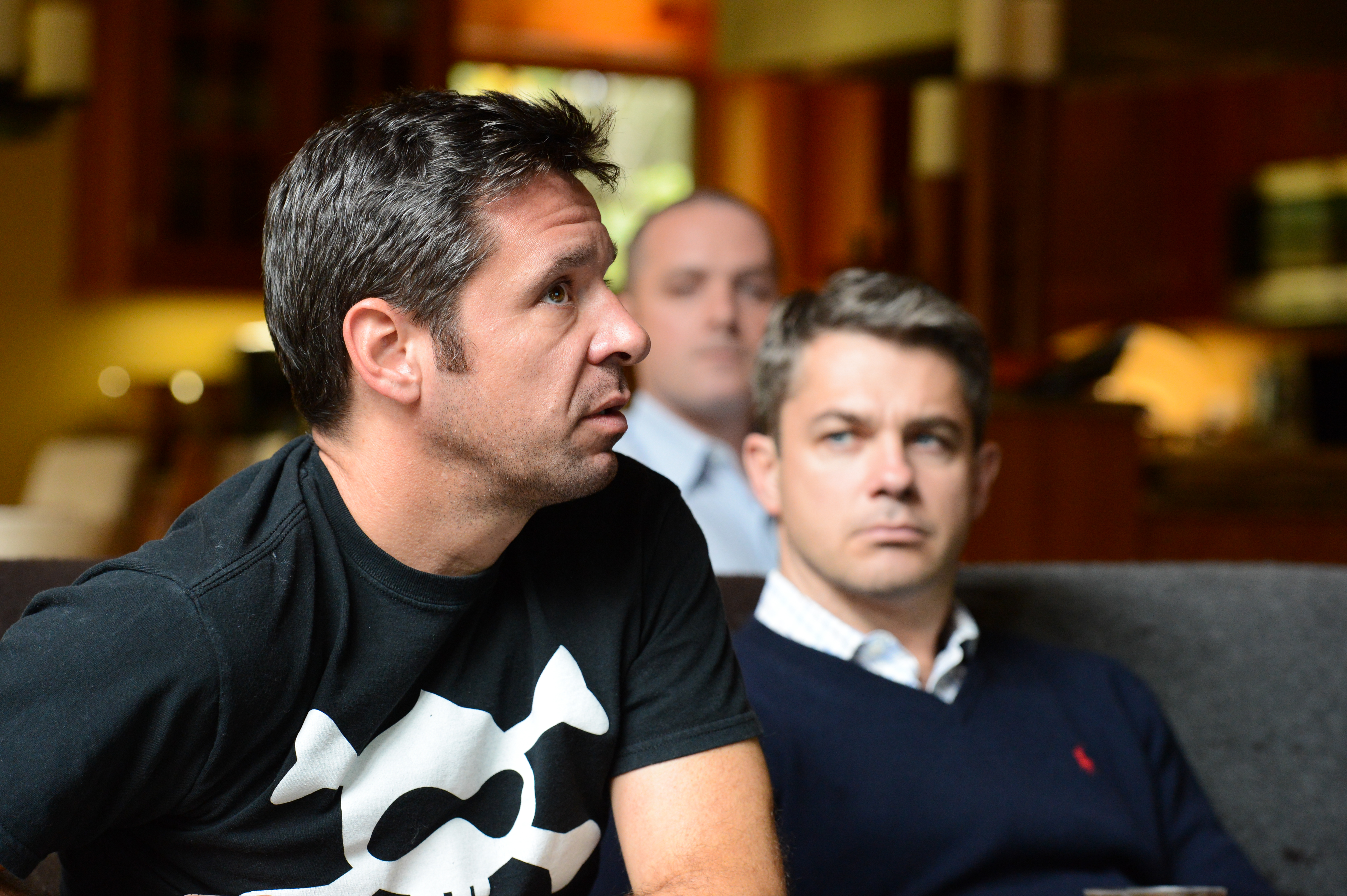 Three men sit indoors, attentively listening to something off-camera.