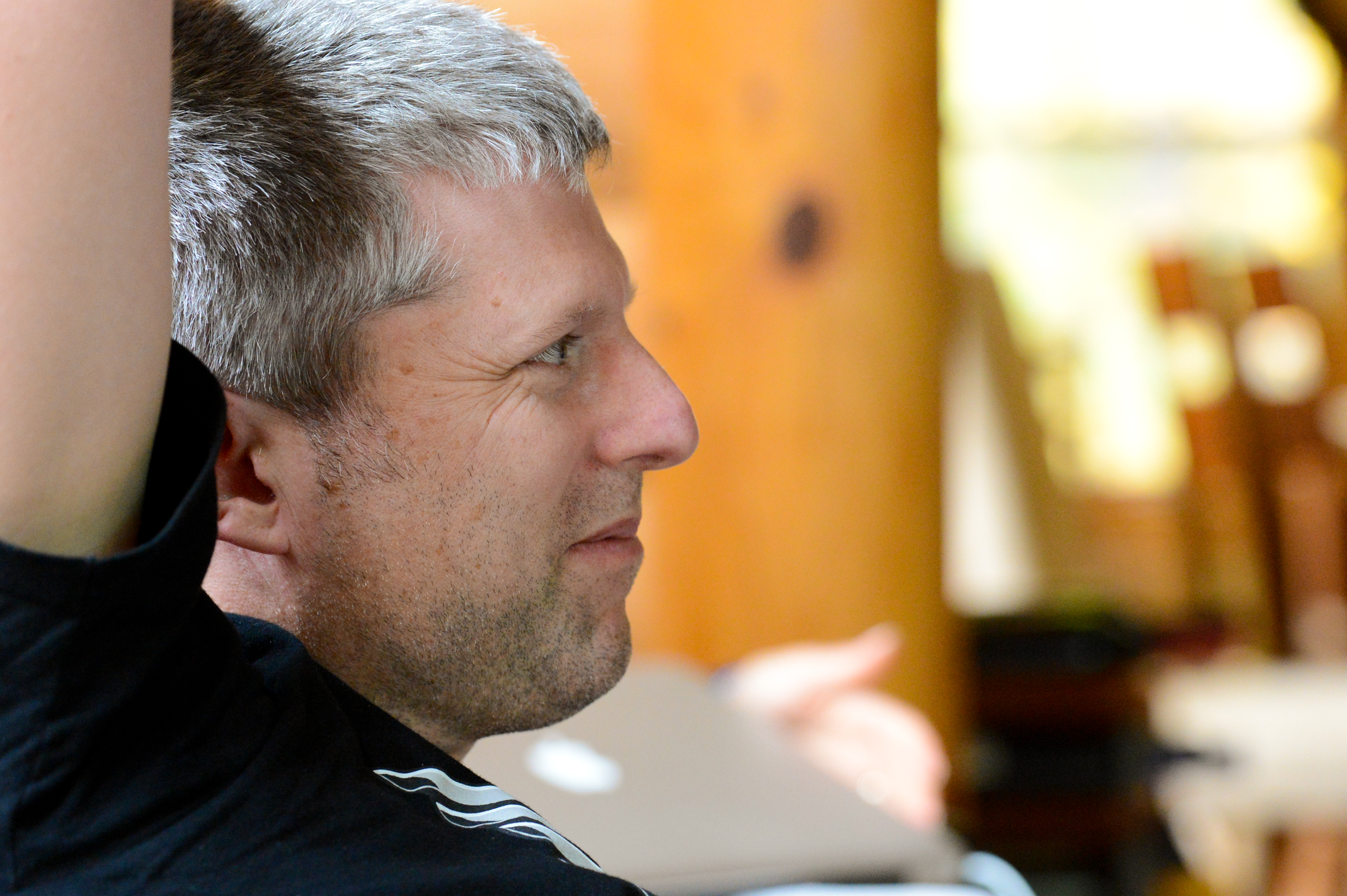 A man with short gray hair raises his arm while looking to the side in a casual setting.