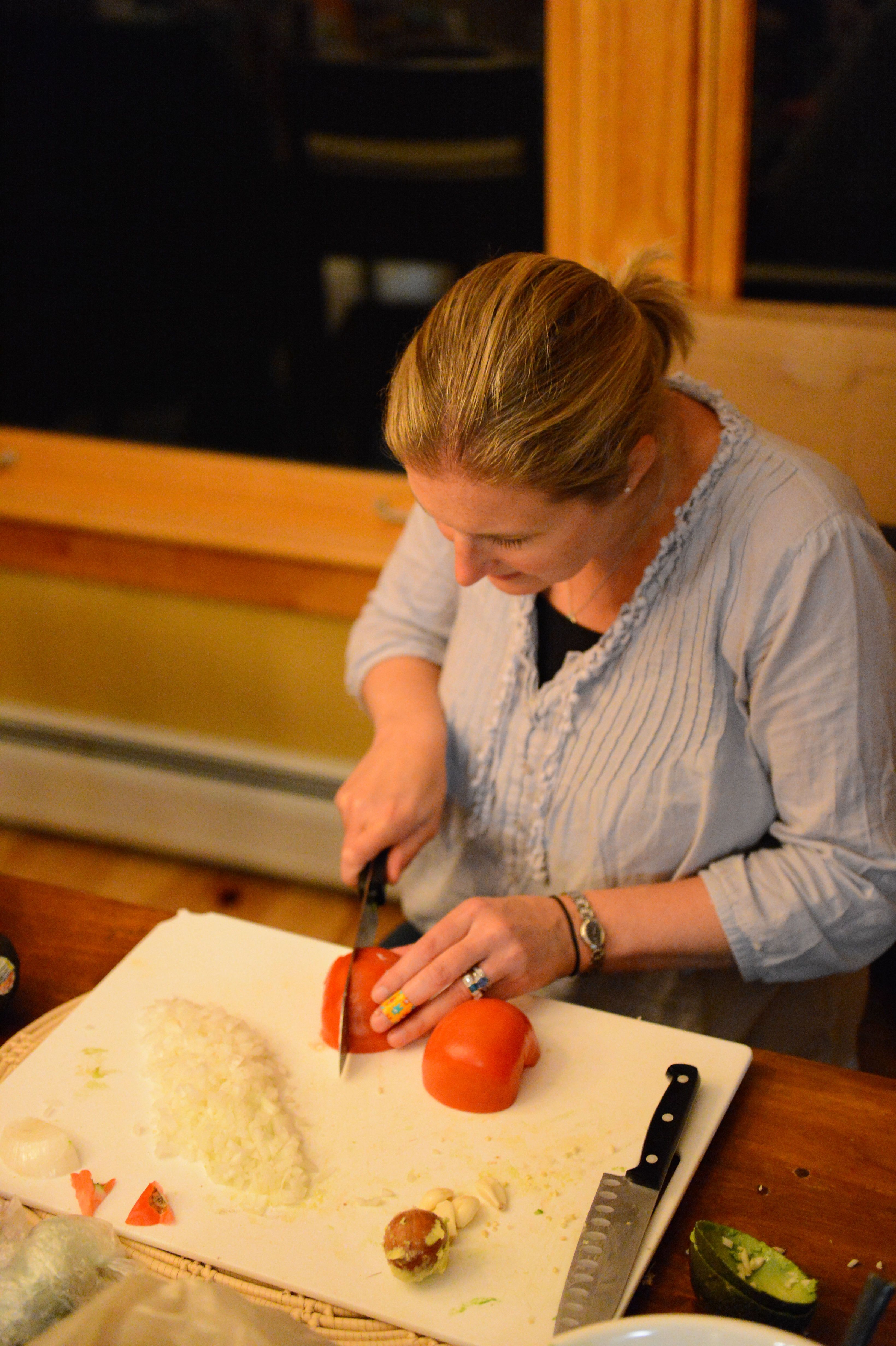 A woman chops tomatoes on a cutting board with diced onions and other ingredients nearby.