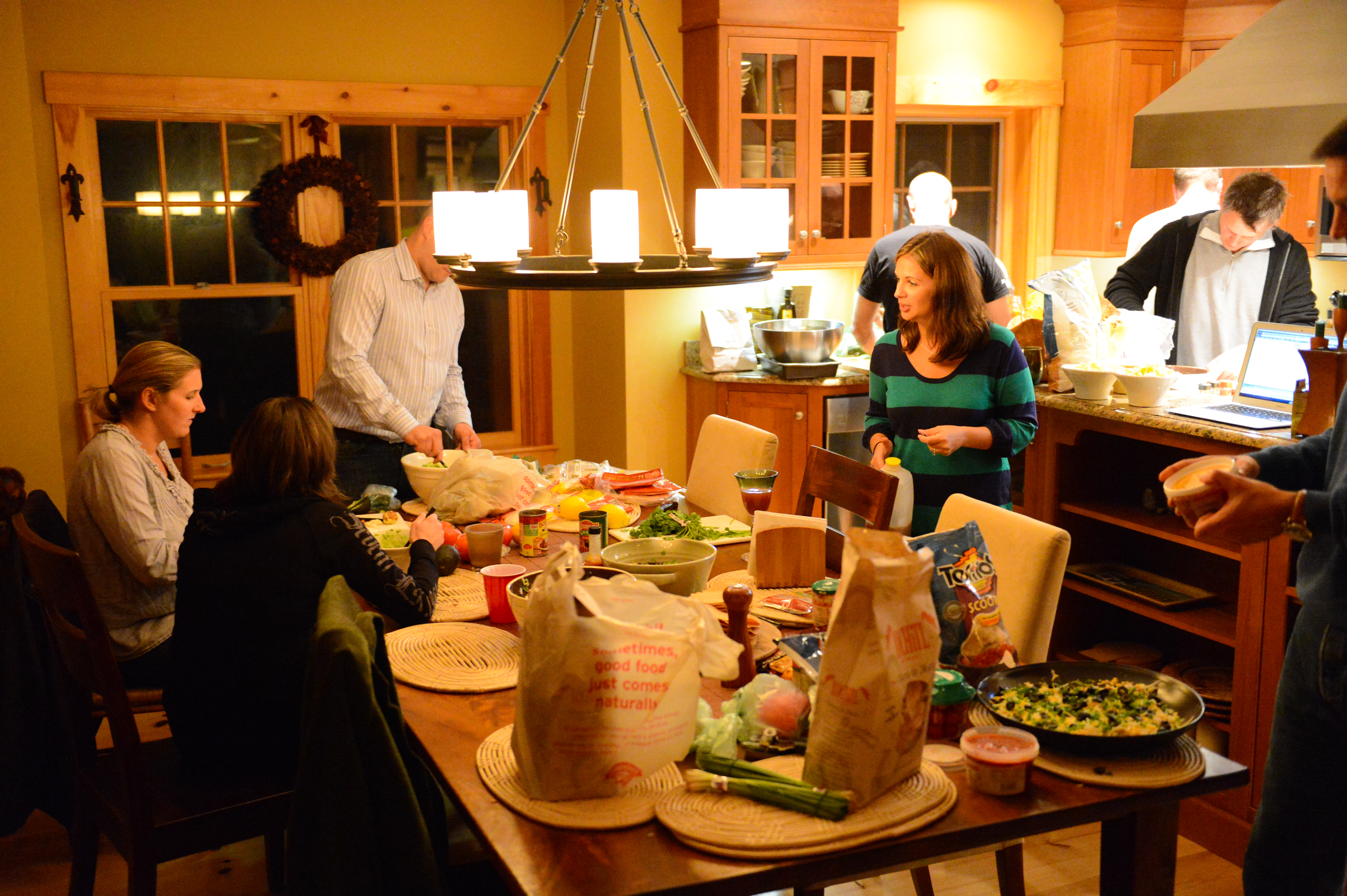 A group of people gathers in a kitchen, preparing and sharing food around a table filled with ingredients.