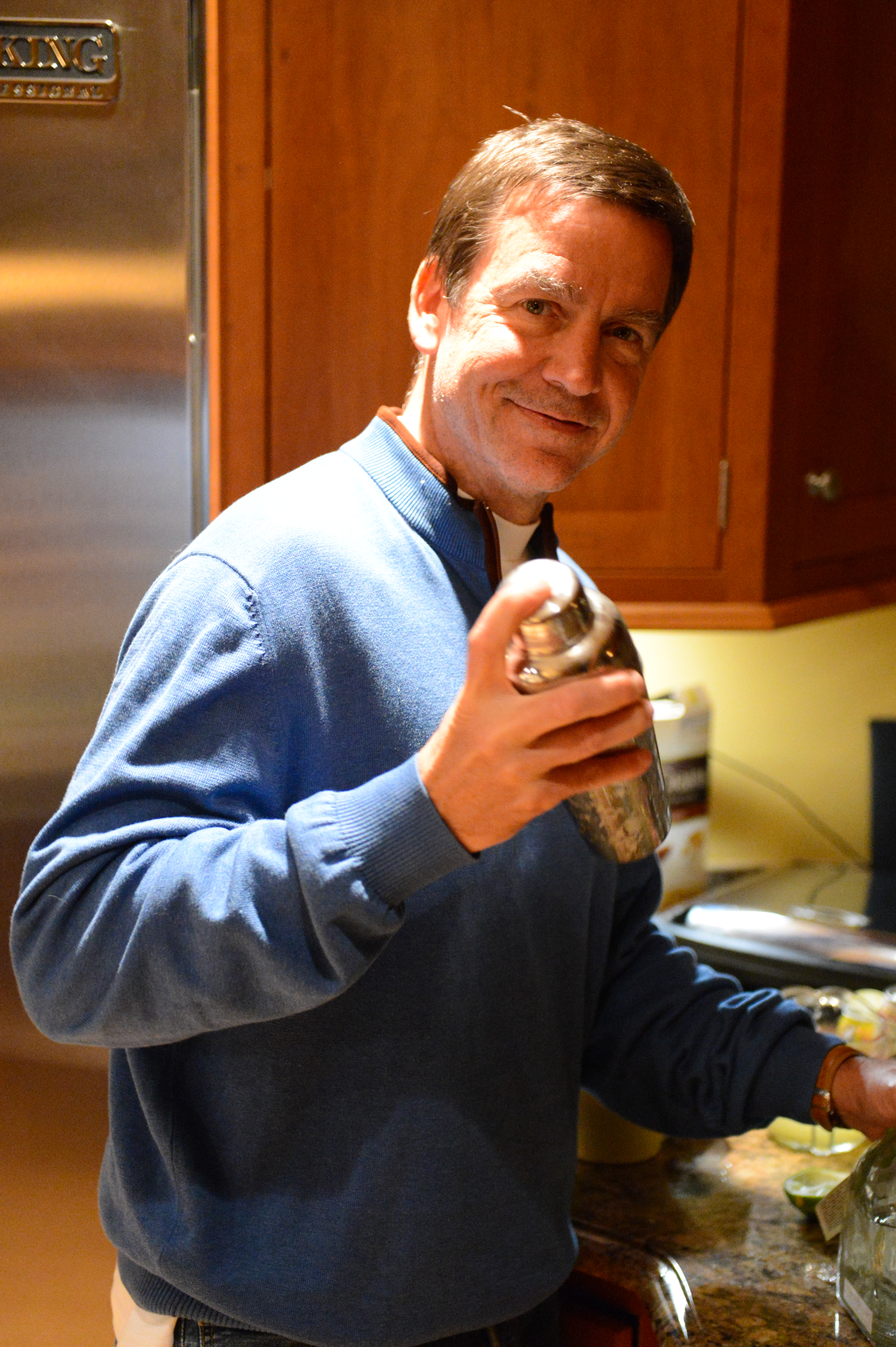 A man in a blue sweater smiles while shaking a cocktail shaker in a kitchen.